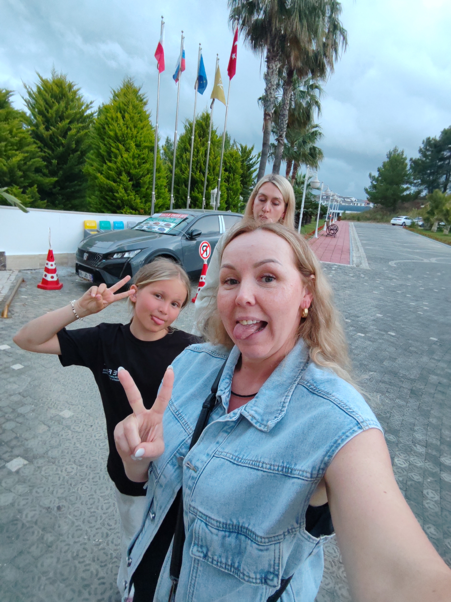 Three women are posing for a selfie on a paved area in Alanya, Turkey. A young girl on the left is sticking out her tongue and making a peace sign. A woman in the foreground is sticking out her tongue. A third woman is standing behind them, looking slightly upward. Behind them is a gray car with advertisements on its hood, flanked by orange and white traffic cones. A row of flags are displayed behind the car, along with trees and some palm trees. A sidewalk is on the right. The sky is overcast.