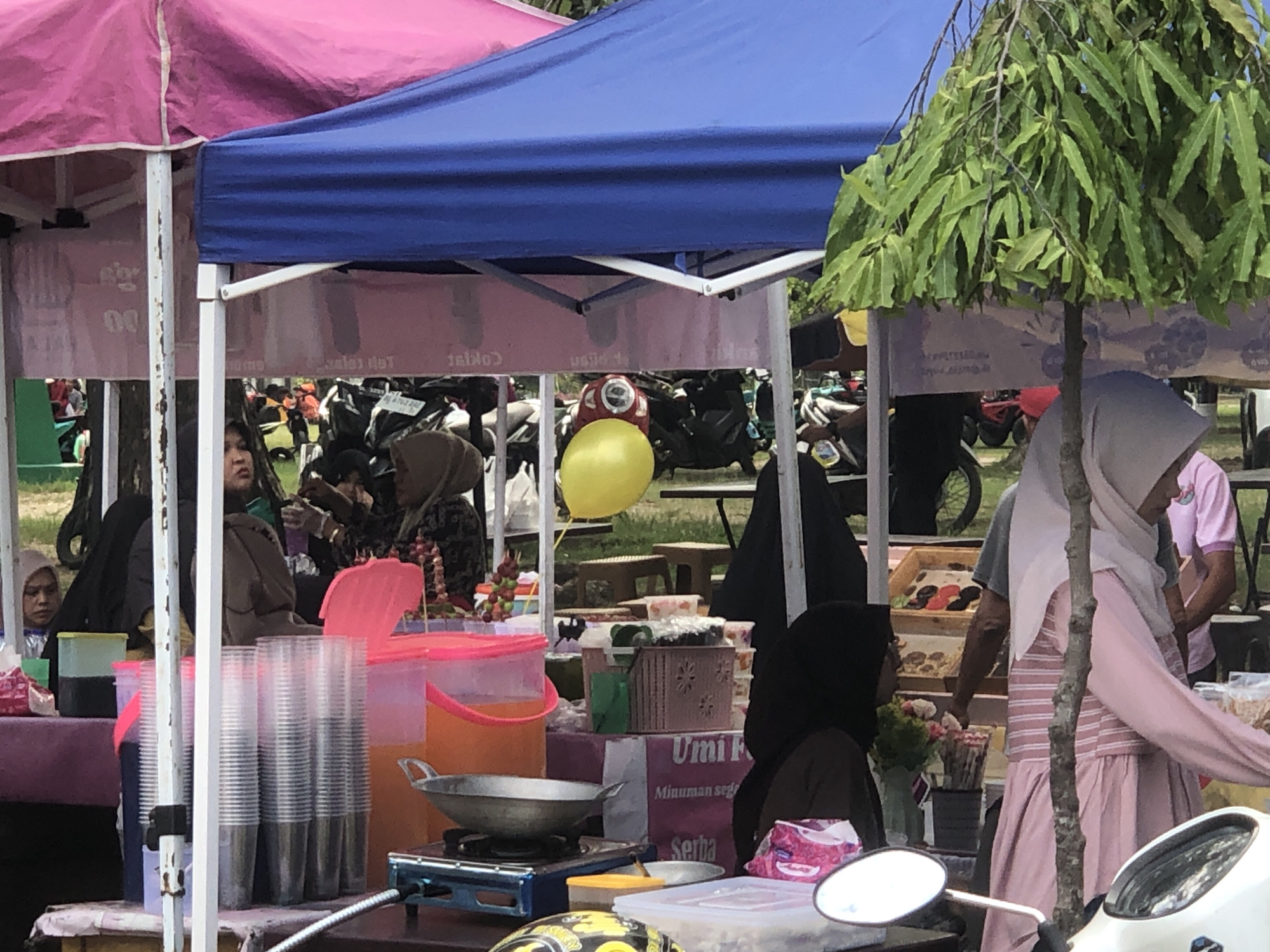 In Lhokseumawe, Indonesia, an outdoor marketplace is bustling with activity. Two adjacent stalls are visible: one under a pink canopy and the other under a blue one. People of various ages are present, some wearing hijabs. A yellow balloon floats behind one stall. Motorbikes are parked in the background. The stalls are set up with food and beverages for sale. A tree partially obscures one of the stalls. There are plastic cups and containers filled with drinks on the tables. Cooking equipment, like a wok on a burner, is also visible.