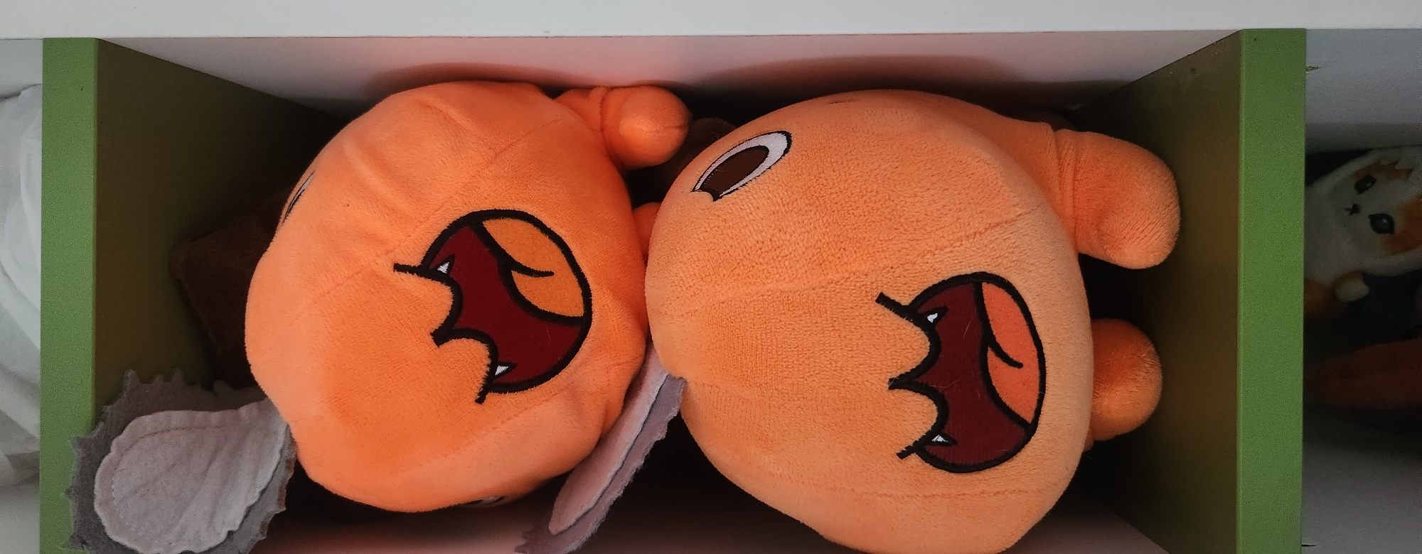 Two plush toys are stacked vertically inside a shelf. The toys are orange and resemble cartoonish characters with wide mouths, sharp teeth, and dark outlines. The top toy has gray ear-like protrusions visible on the left side. Both toys are plump and have rounded features. The shelf appears to be made of wood with a green front edge and a white interior. The background is blurred and out of focus, suggesting an indoor environment. The image is taken from a slightly high angle, looking down into the shelf. The lighting is even, with no harsh shadows. The overall scene depicts a collection of stuffed toys stored on a shelf.