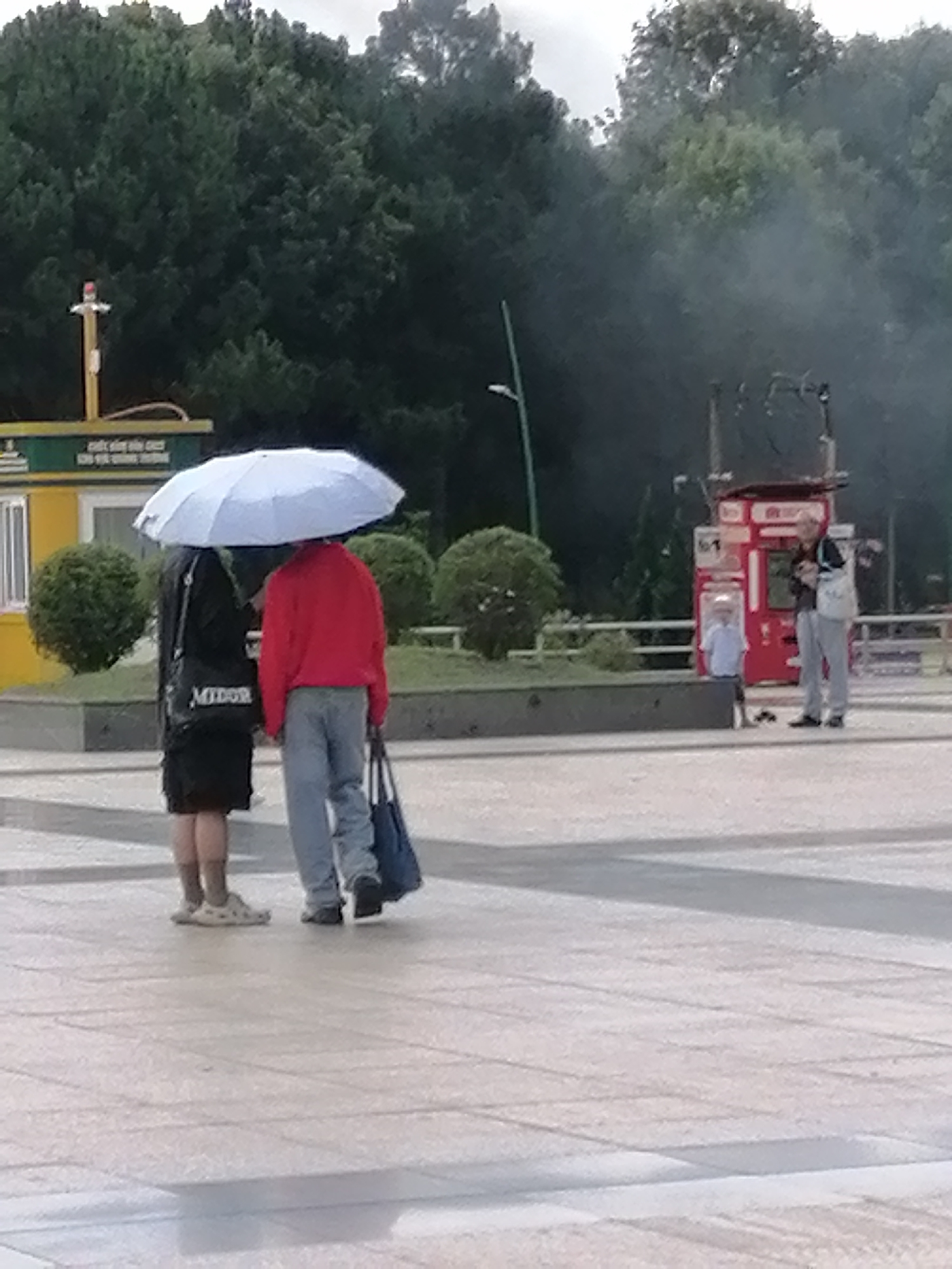 In a paved public square in Da Lat, Vietnam, two individuals stand under a large, light-colored umbrella, shielding them from the elements. The person on the left wears a black outfit and carries a black bag with the word "MINOR" visible. The person on the right is dressed in a red jacket and grey pants, and is holding a dark blue bag. Behind them, a small child in a light blue shirt stands near a red vending machine, with an adult observing from a distance. The background features lush green trees and a cloudy sky, suggesting overcast weather. The scene is captured from a medium distance, with the focus on the people and their immediate surroundings.