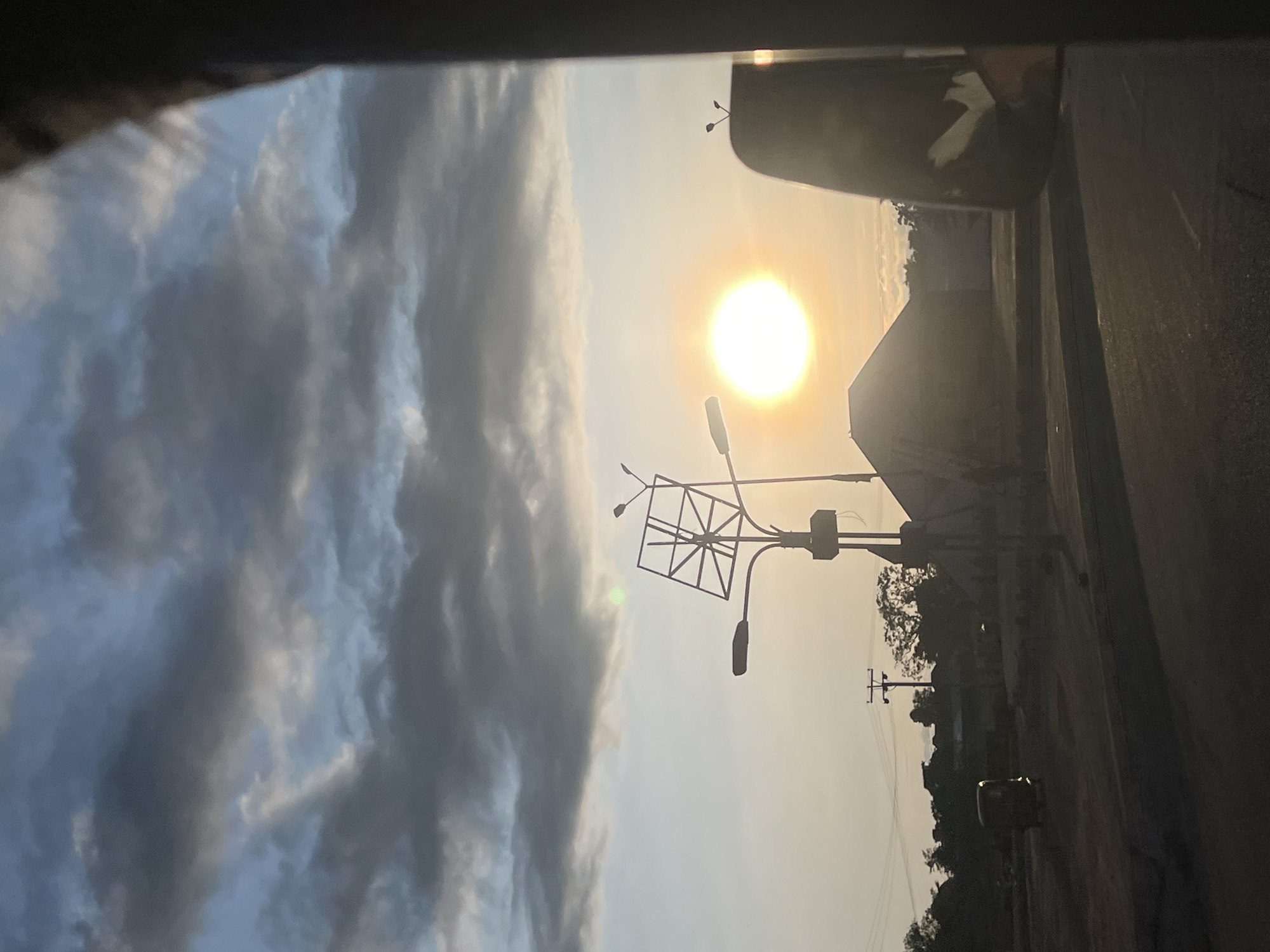 This image, captured in Jalingo, Nigeria, depicts an outdoor scene during either sunrise or sunset. The upper portion of the sky is dominated by textured grey and blue-grey clouds, while the lower sky transitions to a clear, bright orange-yellow hue near the horizon. A prominent, intensely luminous, circular white-yellow sun is positioned low on the right side of the frame.
In the mid-ground, a tall utility pole is centrally located, supporting two streetlights and a distinctive, open-lattice metal framework, which appears to be designed for a billboard or a solar panel array. To the left of this pole, a dark, three-wheeled vehicle, consistent with an auto-rickshaw common in Nigeria, is visible on an unpaved road surface, exhibiting minor light reflection. Further in the mid-ground, multiple power lines and wooden utility poles are silhouetted against the sky. Various dark, indistinct buildings, including one with a peaked roof, along with scattered silhouetted vegetation, are visible across the mid-ground. The immediate foreground consists of a dark road surface. A dark, vertical obstruction on the far right, encompassing a partial view of a vehicle's side mirror, indicates the photograph was taken from inside a vehicle. No explicit human figures are discernible.