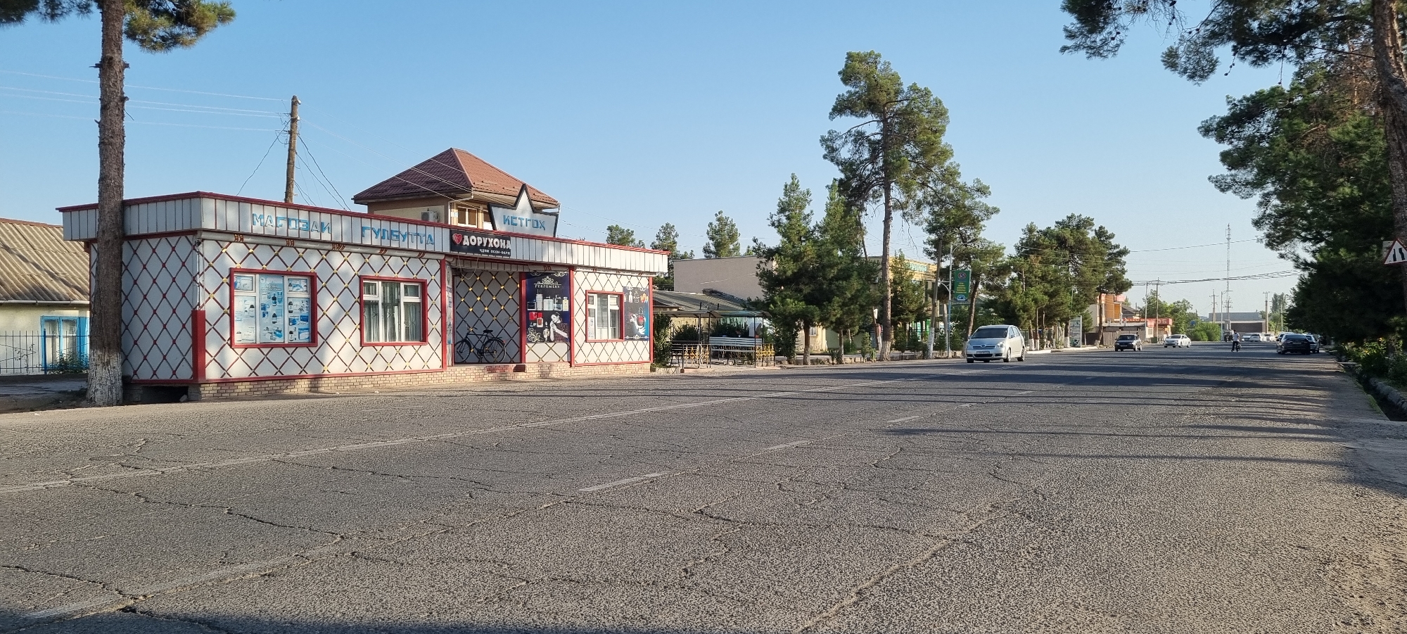 The image shows a street scene in Ismoili Somoni, Tajikistan, during daylight. A small, white and red patterned building, possibly a shop or kiosk, is prominently featured on the left side of the road. The building has signage in a Cyrillic script, with "МАГАЗИН" (Magazin - Shop) visible at the top, and further text including "ГУЛЕУТТА" and "ДОРУХОНА" (Doruxona - Pharmacy) above different sections of the building. It has multiple windows displaying products, including one showing what appears to be toiletries or cosmetics, and another with various bottles. A bicycle is parked inside the shop. The building is situated near a paved road with lane markings. Several vehicles are visible on the road, including a silver minivan driving towards the viewer and other cars and a motorcycle further down the street. Tall trees line the street on both sides, and the sky is clear and blue. The environment appears to be a semi-urban or suburban area with residential buildings and other commercial structures visible in the background.