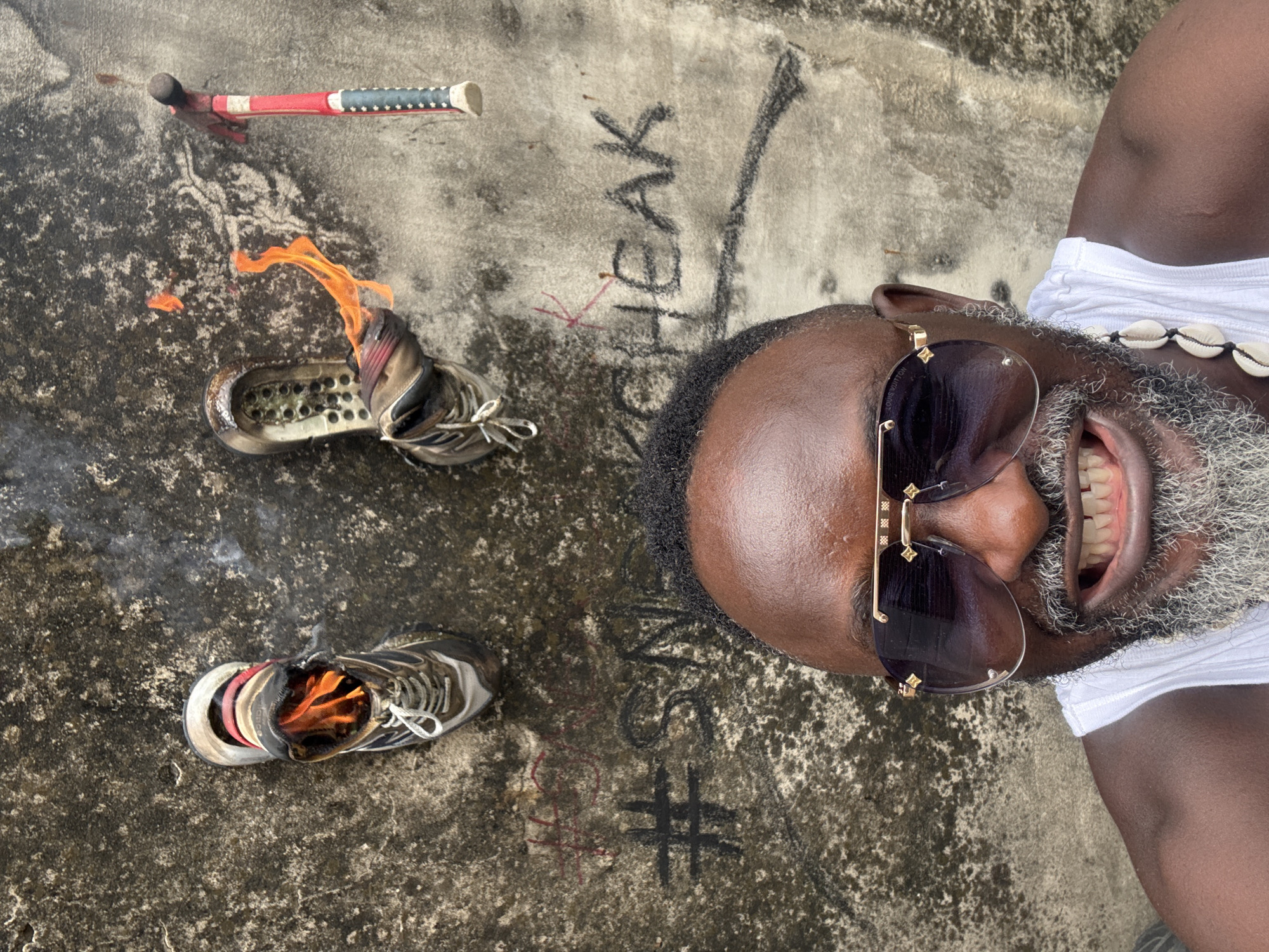 The image depicts a selfie of an adult male in the city of Ado, Nigeria, positioned against a weathered concrete wall. The man, with dark skin, a short dark afro hairstyle, and a prominent gray beard, wears large, dark aviator sunglasses with visible "Louis Vuitton" branding on the frame and a white ribbed tank top. A necklace of white cowrie shells is visible on his right shoulder. He displays a wide, toothy grin, revealing yellowed teeth.
Above his head, two athletic sneakers are affixed to the textured, discolored wall. The sneaker on the left is upright, with visible flames originating from its interior. The sneaker on the right is positioned horizontally with its sole facing outwards, also exhibiting significant flames and smoke emanating from its upper. To the upper right, a tool with a red and dark blue handle, resembling a pickaxe or hammer, is attached to the wall. Below the burning sneakers, the phrase "#SNEAKCHEAK" is written in dark graffiti on the wall, accompanied by additional faint red markings above it. The wall's surface is rough, concrete-like, and shows extensive discoloration and dark patches.