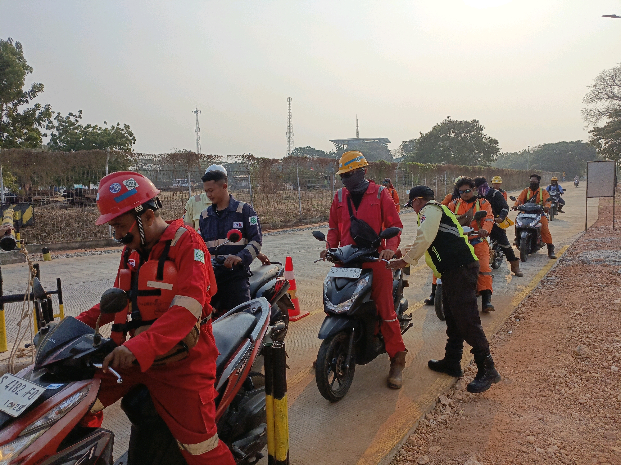 The image depicts a group of workers in a construction or industrial setting in Jenu, Indonesia. The workers are predominantly wearing red or orange safety suits and helmets, indicating they are likely engaged in hazardous work. Several individuals are positioned around motorcycles, suggesting they are either preparing to depart or have recently arrived at the location.
In the foreground, a worker in a red suit with a red helmet is seated on a red motorcycle, holding the handlebars. To their left, another motorcycle is partially visible, with a worker in a red suit and red helmet standing near it. Behind the first seated worker, a man in a dark blue uniform and a white helmet is looking down.
Further back in the scene, a line of workers on motorcycles is visible. One person in a red suit and yellow helmet is on a black motorcycle. To their right, another worker in an orange suit and yellow helmet is on a motorcycle, wearing sunglasses. More workers on motorcycles and individuals in various colored safety gear are spread throughout the background, moving away from the viewer.
The environment appears to be an outdoor, unpaved area with some construction barriers and fencing visible. In the distance, there are trees and what appear to be communication towers. The overall lighting suggests it is daytime, possibly early morning or late afternoon, given the warm tone of the light. The ground is covered in reddish-brown gravel and dirt, characteristic of an industrial or construction site.