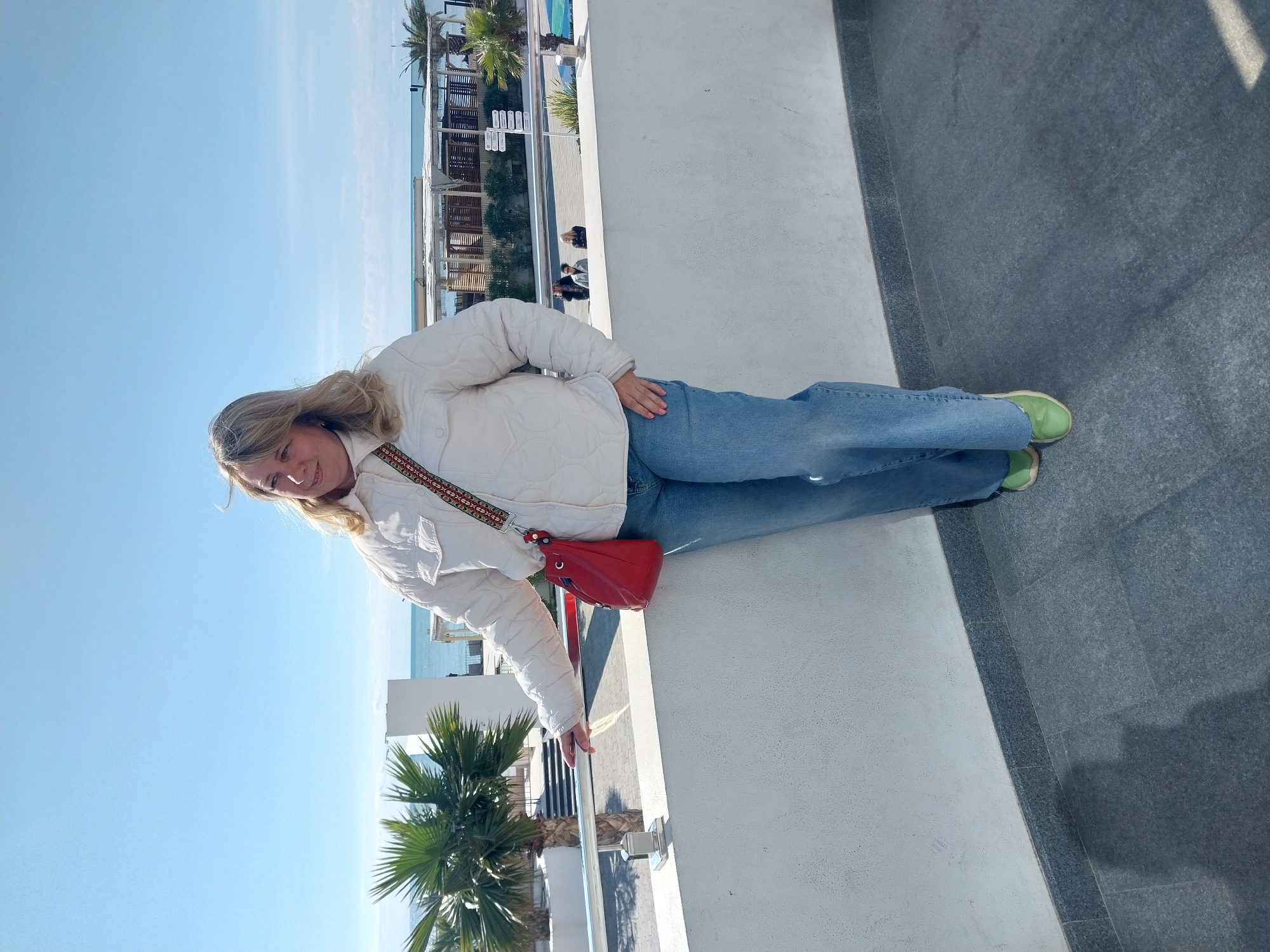 The image shows a woman standing on a white concrete ledge with a darker gray concrete area to her right. She is wearing a white quilted jacket, blue jeans, and light green sneakers. A red crossbody bag hangs on her left side. The background features a clear blue sky and a view of the sea. In the distance, there are buildings and palm trees. Based on the architecture and palm trees, the location is likely in a coastal area, possibly in Sochi, Russia.