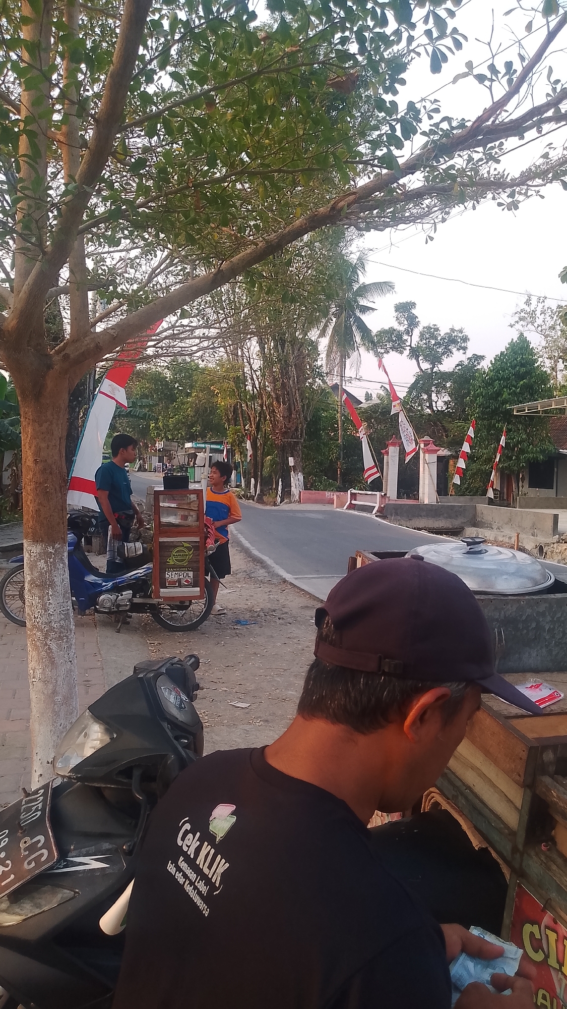 The image depicts a street scene in Karanglangu, Indonesia, with a focus on two young men and a motorcycle. A man in a blue t-shirt is standing next to a blue motorcycle, facing a younger man in an orange and blue shirt. The younger man is standing next to a mobile food cart that is attached to the motorcycle. The food cart has various items displayed on it, including bottles and packaged goods. The younger man appears to be interacting with the food cart. In the foreground, another person wearing a dark t-shirt and a baseball cap is partially visible, with their back to the camera, and they are holding what appears to be money. The street is paved, and there are trees and flags lining the road. In the background, there are houses and more trees. The lighting suggests it is either early morning or late afternoon.