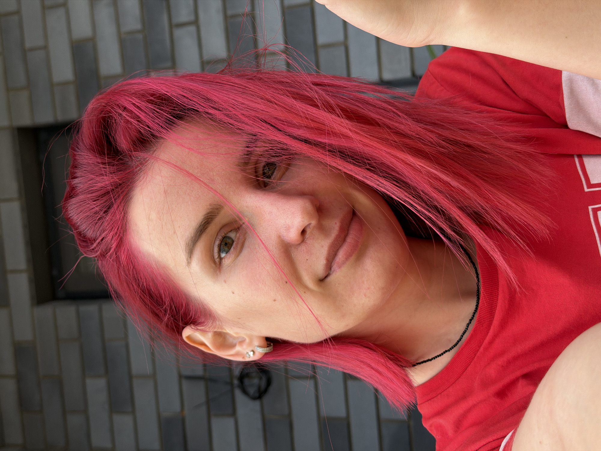 A young woman with bright pink hair is captured in a close-up, selfie-style photograph. She is wearing a red t-shirt and a thin black beaded necklace. Her left ear is adorned with two small stud earrings. The background appears to be a grey brick wall, possibly a building exterior. The lighting suggests it is daytime. The image is a medium shot, focusing on the woman's face and upper torso. No specific location in Chaikovsky, Russia, is identifiable from the visual information present in the image.