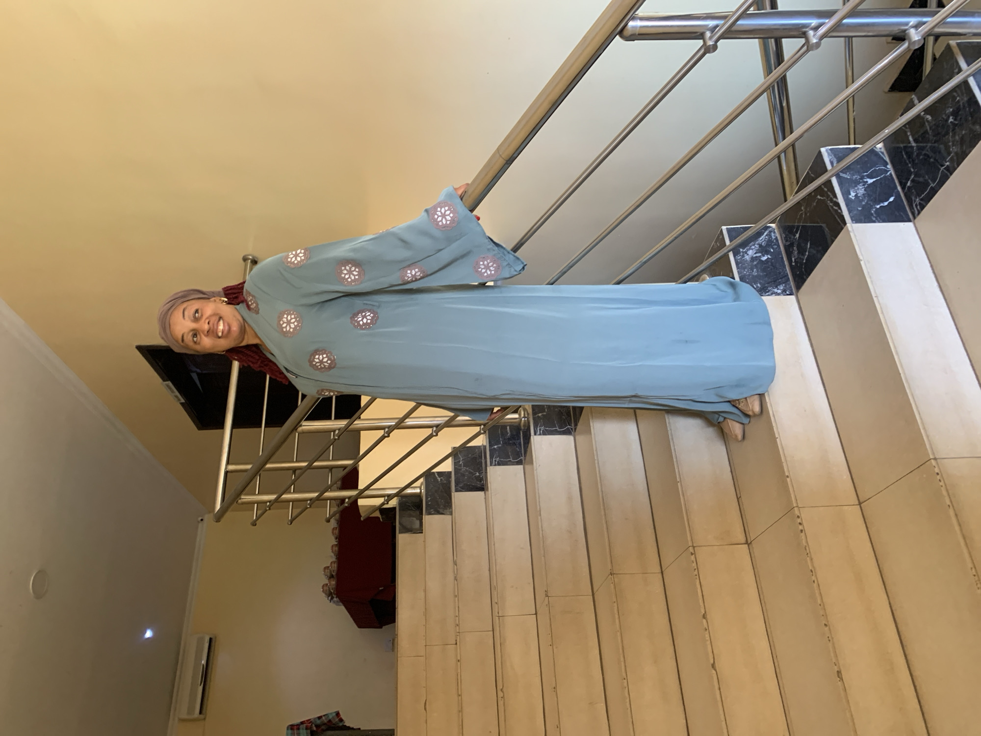 A woman stands on a staircase, dressed in a long, blue, decorated garment. The staircase has a stainless steel railing. Black and white tile trim is visible along the side of the stairs. The woman is smiling. The location is presumably an interior staircase in a building in Gura Top, Nigeria.