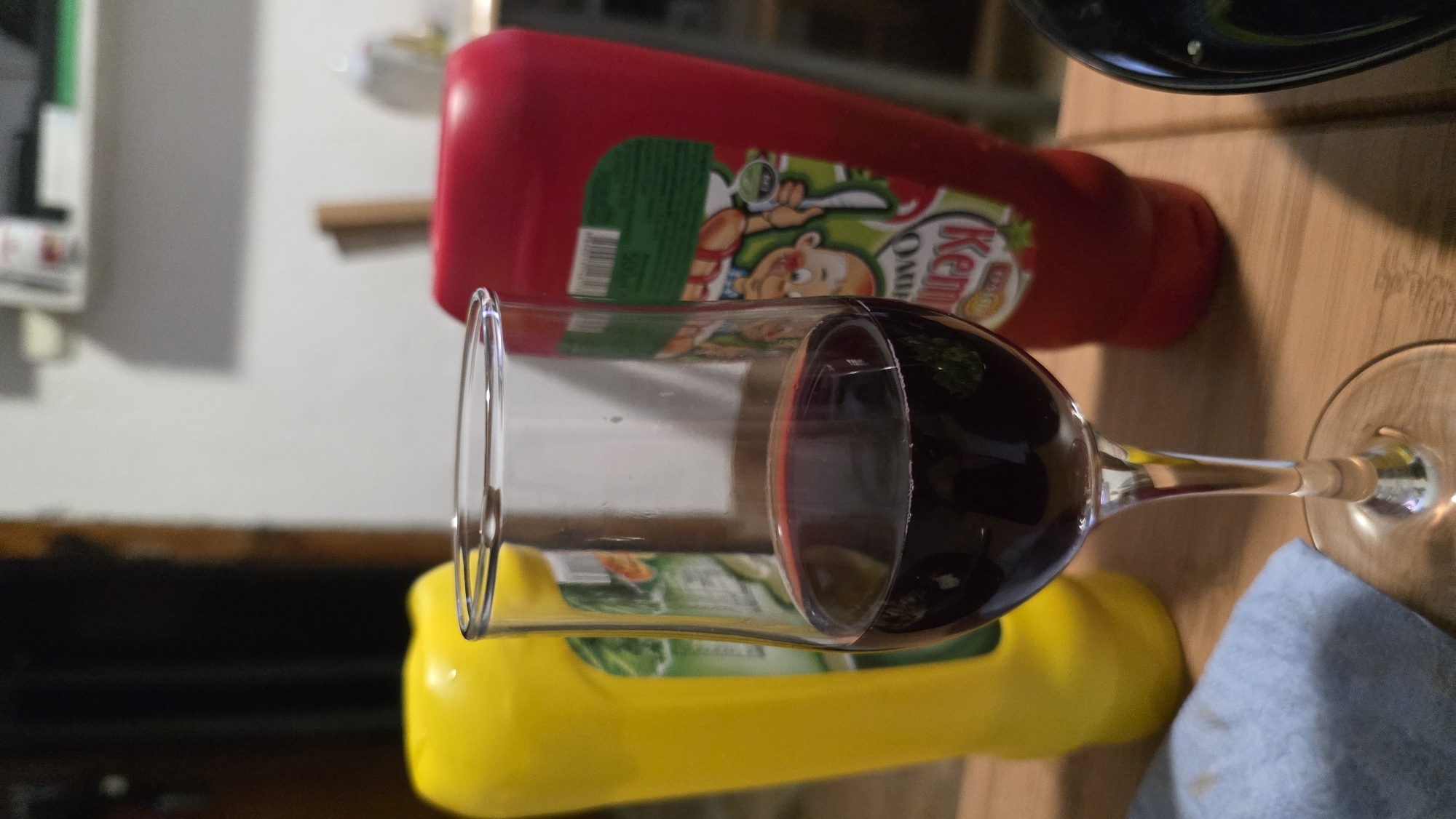 A wine glass filled with red wine is on a wooden surface next to bottles of ketchup and mustard. The label on the ketchup bottle is partially visible, showing a cartoon character and the word "Kem". The bottles are oriented horizontally in the image. A blue cloth is near the base of the wine glass. The exact location within Blagoevgrad, Bulgaria is not discernible from the image.