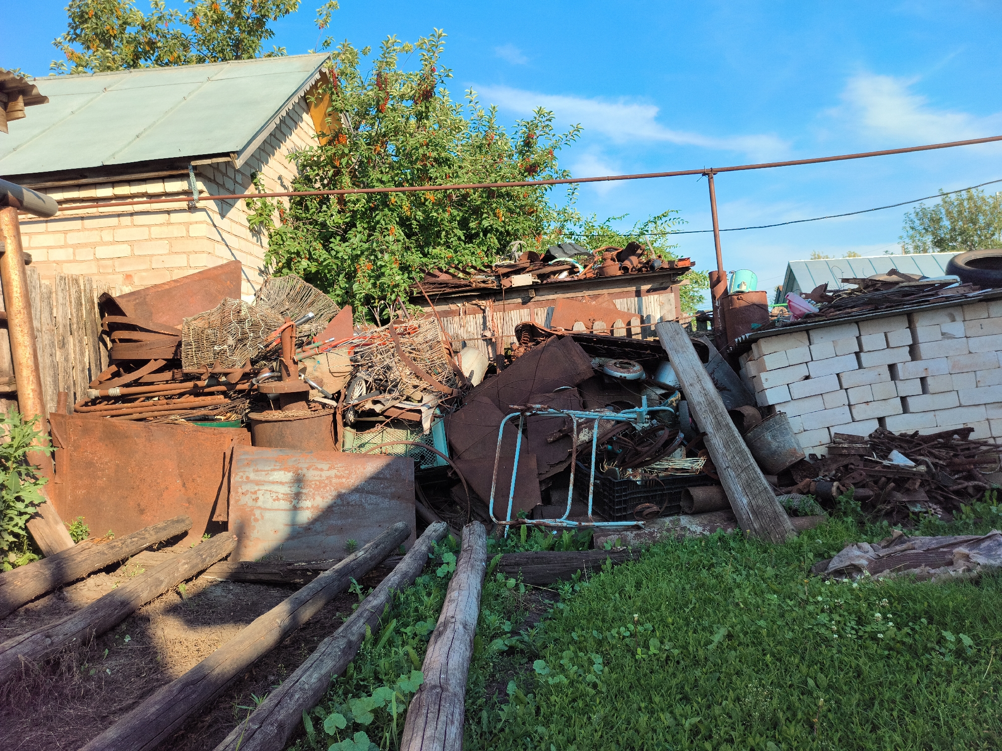 The image depicts a cluttered outdoor scene in Алькино, Russia, likely a yard or storage area. A large pile of scrap metal and other discarded items is the central subject. The materials include rusted barrels, sheets of metal, wire cages, wooden planks, and what appears to be parts of a bicycle or cart. In the background, a light-colored brick building with a green metal roof is visible, partially obscured by a lush green tree laden with red berries. Further in the distance, a white brick wall with more piled-up debris is present. The ground is covered with dry earth and patches of green grass. Several weathered wooden logs are placed horizontally on the ground in the foreground.
The time of day appears to be late afternoon or early evening, given the strong shadows cast by the objects and the warm sunlight. The weather is clear and sunny, with a bright blue sky and a few white clouds. There are no people visible in the image. The overall impression is one of disarray and neglect, suggesting a place where items are stored or accumulated for later disposal or recycling. No text is visible in the image.