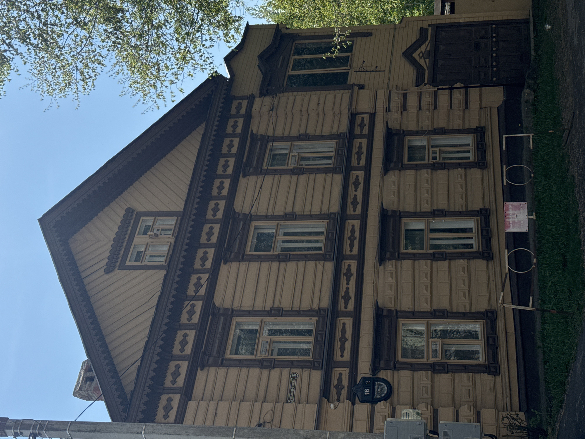 The image captures a three-story wooden building in Kazan, Russia, characterized by its intricate ornamental carvings around the windows and along the roofline. The building's facade is predominantly brown and tan, with darker brown accents highlighting the decorative elements. The windows are rectangular and evenly spaced across the facade. A sign displaying the number "16" and the street name in Russian is visible near the lower left corner of the building. To the right of the building is a grassy area with a metal post and hoop. Above the building, branches and leaves from trees are visible against a clear blue sky.