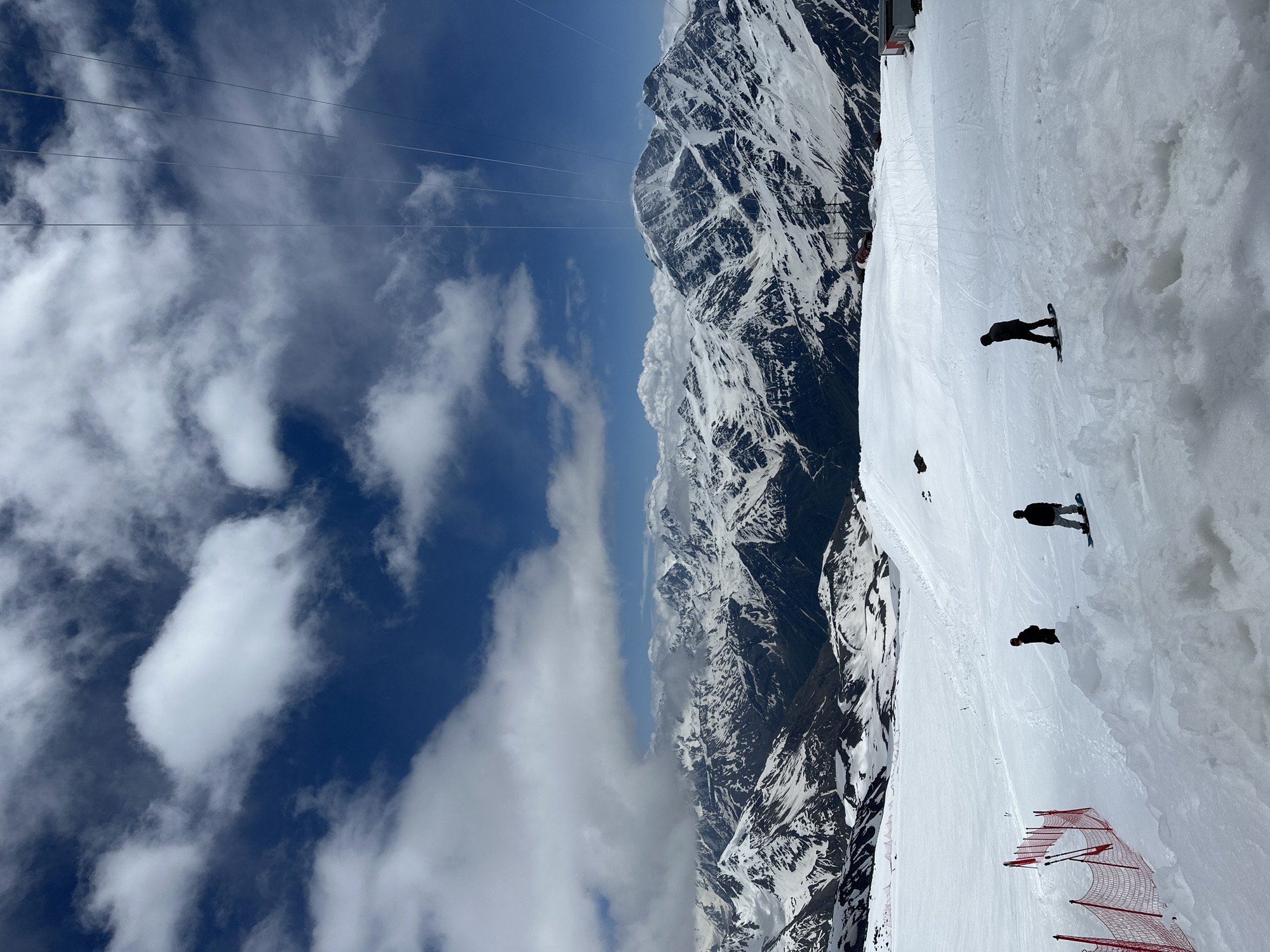 The image captures a breathtaking mountain vista, likely the ski resort of Terskol, Russia, given the context of the surrounding snowy peaks and ski infrastructure. In the foreground, a vast expanse of white snow covers the slope, marked by tracks from skiers and snowboarders. Three figures are visible on snowboards, silhouetted against the bright snow, each engaged in the activity. To the left, a red safety net with poles delineates a section of the slope.
In the midground, the snow-covered terrain continues, with a gentle slope leading towards the majestic mountain range that dominates the background. These mountains are dramatically rugged, their peaks covered in snow and shadows, suggesting their immense scale and altitude.
The sky above is a brilliant blue, dotted with fluffy white clouds that cast subtle shadows on the mountains. A visible ski lift cable stretches across the upper portion of the frame, hinting at the resort's operational capacity. The overall scene evokes a sense of a crisp, clear day at a high-altitude ski resort, with the raw beauty of the natural landscape as the primary focus.