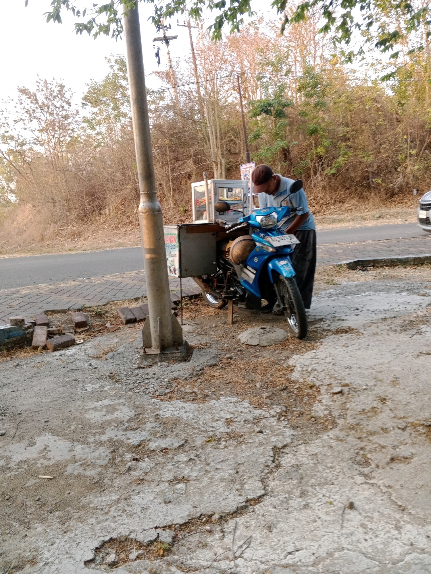 An Indonesian man wearing a brown cap and a light blue shirt stands by a blue Yamaha motorcycle in what appears to be a roadside setting. The motorcycle has a sidecar-like storage compartment. A food cart with glass panels is attached to the motorcycle. The man is bent over, appearing to be working on or interacting with the motorcycle. The scene is outdoors, with a dry, arid landscape of trees and vegetation in the background. The ground in the foreground is rough, with patches of concrete and dirt. A paved walkway runs alongside the road in the mid-ground. The lighting suggests it is daytime, possibly late afternoon due to the warm tones. The license plate on the motorcycle is partially visible. The image is taken from a slightly low angle, looking up towards the man and motorcycle.