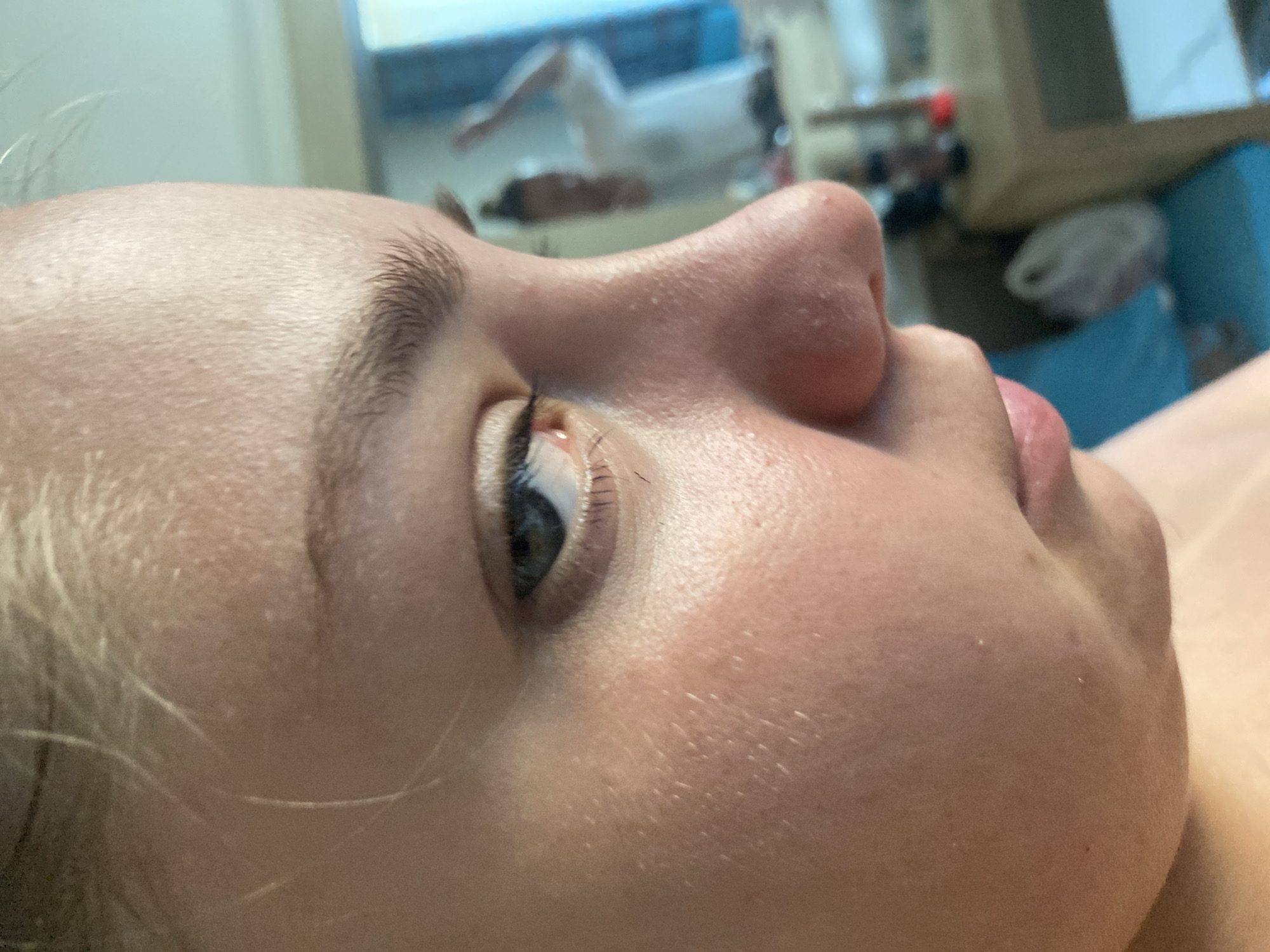 The image is a close-up, profile view of a young woman's face, focusing on her eye, nose, and cheek. Her skin appears fair with some texture. Her left eye is open and looking downwards. Her blonde hair is visible at the top of the frame. In the blurred background, a mirror reflects a person in a white shirt with their arm raised. The setting appears to be indoors, possibly a hotel room or bathroom, with furniture and a blue curtain visible. The image is not located in Nesebar, Bulgaria.