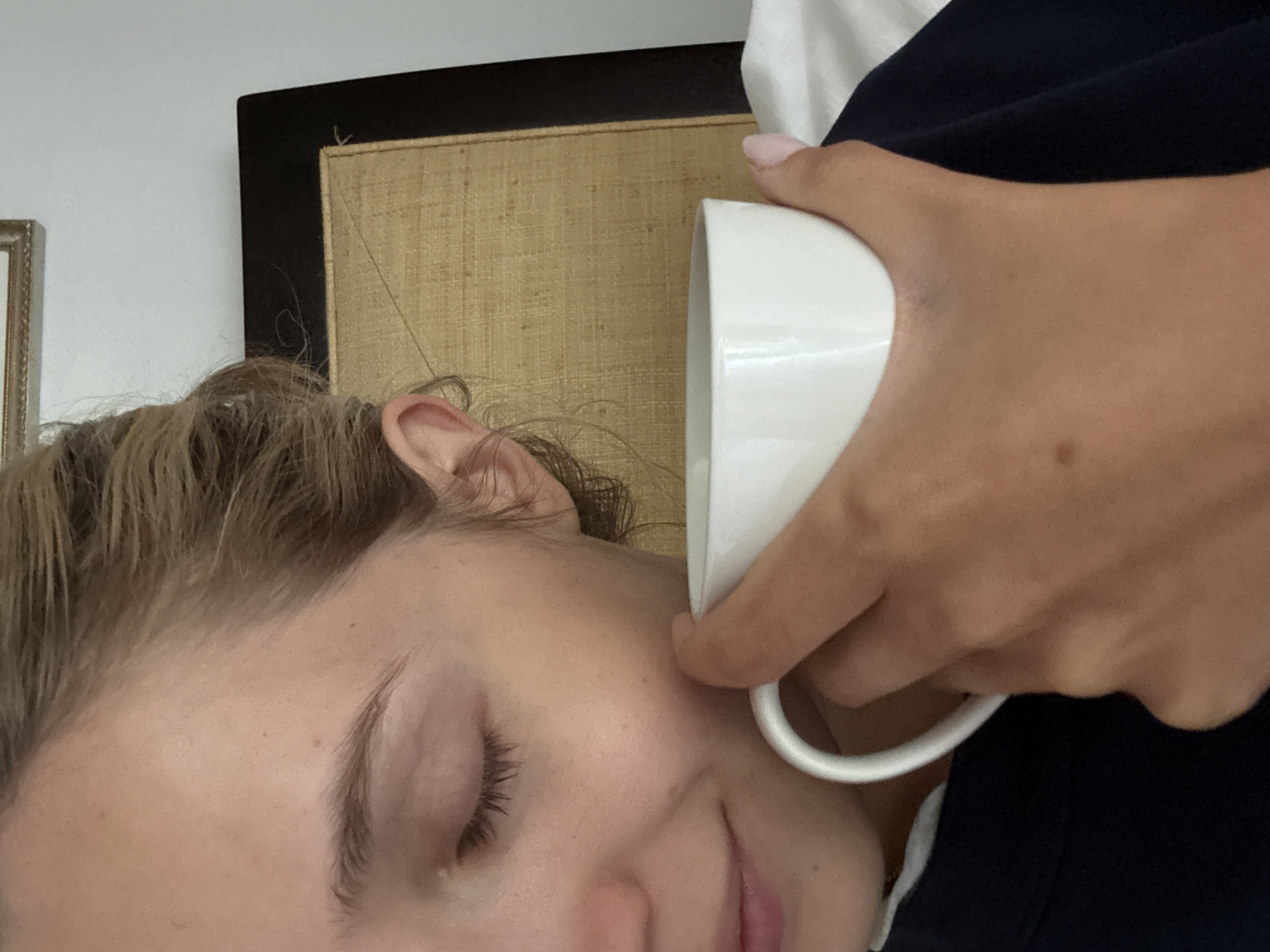 A close-up, top-down shot captures a person's face and hand holding a white mug. The person's eyes are closed, and they appear to be smelling or sipping from the mug, their face partially obscured by it. Their hair is pulled back. The background shows a woven headboard and a white wall, suggesting an indoor domestic setting. The image is devoid of any identifying features that would place it within the city of Ciudad de México, Mexico.