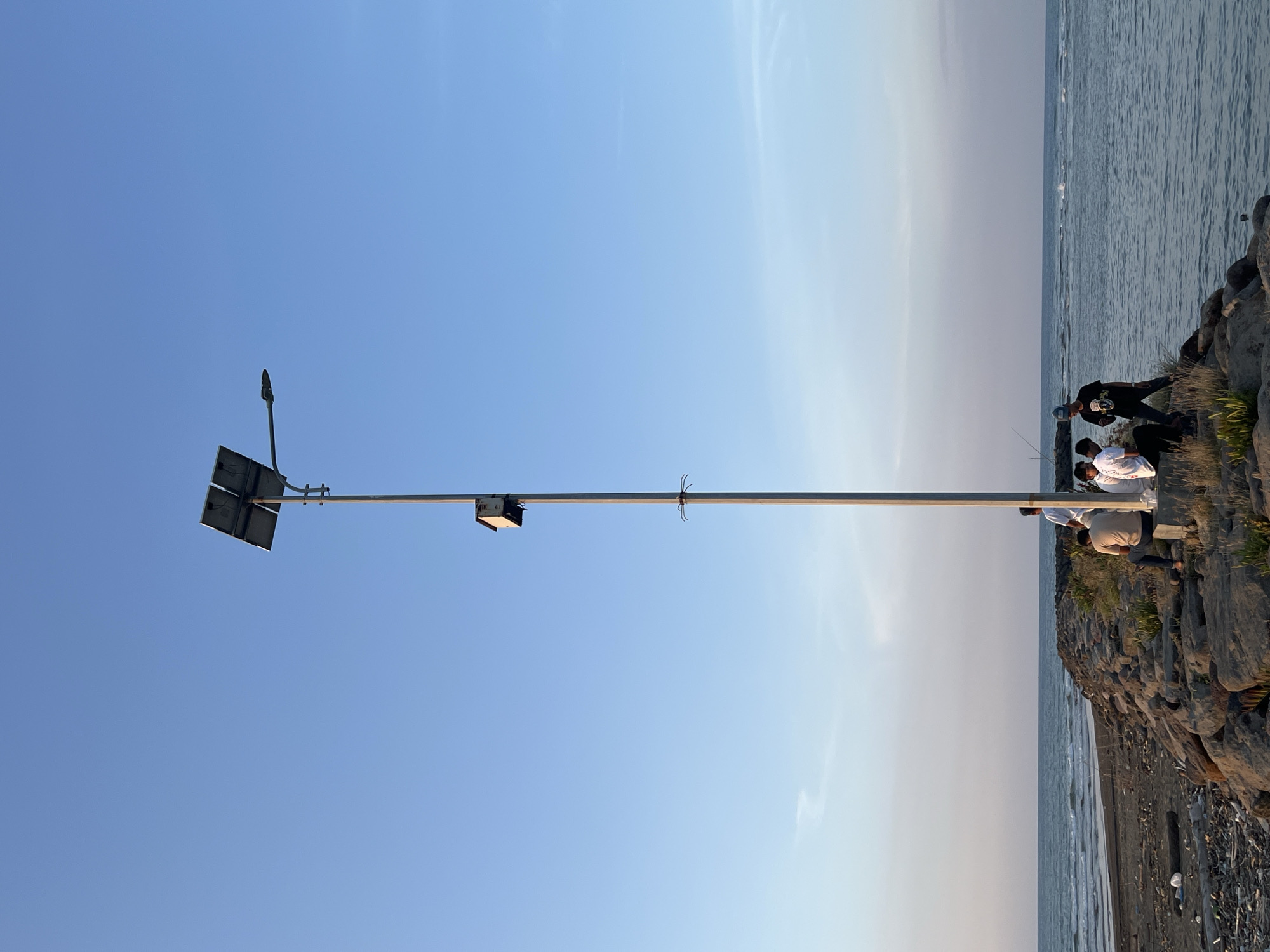 A group of five young men are gathered on a rocky pier that extends into the ocean. The pier is lined with large, dark stones and some sparse vegetation. On the pier stands a tall, slender lamppost powered by solar panels mounted at its top. A small, light-colored box is attached to the lamppost. The men are casually dressed; three are sitting on the stones, and two are standing. One man is holding a fishing rod. The ocean is calm with gentle waves lapping at the shore. The sky above is clear and a pale blue, with a hint of pink and yellow near the horizon, suggesting either sunrise or sunset. The scene appears to be located on the coast of Meunasah Jurong, Indonesia, a coastal village known for its beaches and ocean views.