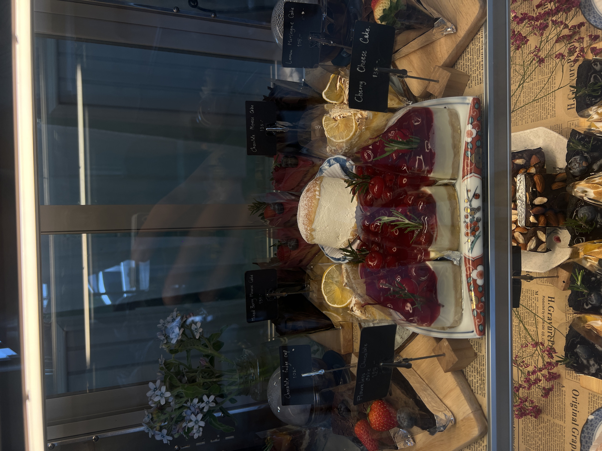 A display case features various desserts, including cakes, with price tags. Among the offerings are "Lemon Meringue Cake," "Cherry Cheese Cake," "Chocolate Mousse Cake," "Mixed Berry Mousse Cake," and "Floureless Chocolate Cake." One item is labeled "Chocolate fudge cup." The scene is likely a bakery or café in the city of Unknown, Unknown. The cakes are displayed on different surfaces including wooden boards and decorative plates. There are also flowers in a vase within the display case.