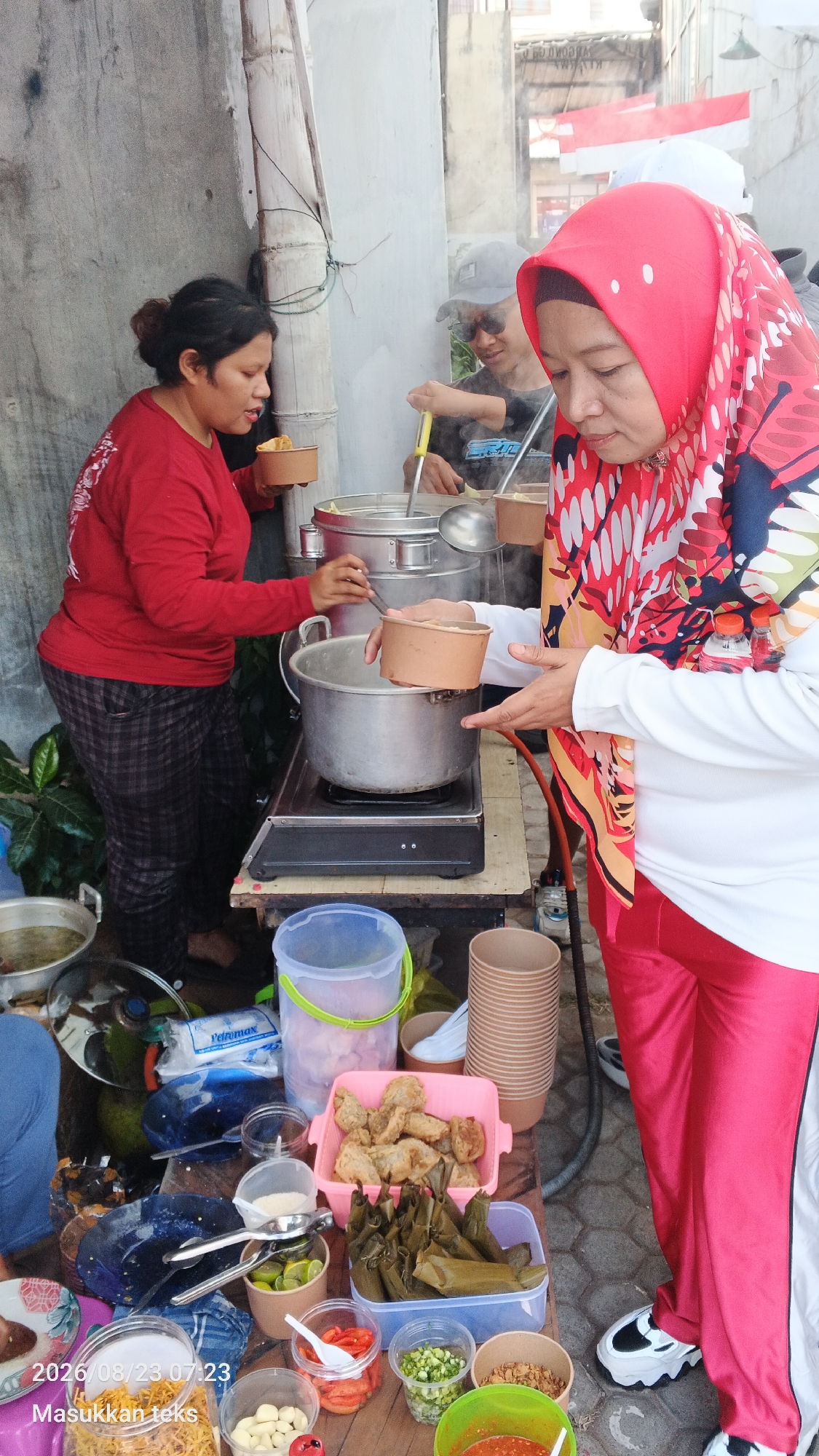 In the city of Malang, Indonesia, at street level, an outdoor food stall is in operation. A woman wearing a red headscarf and red pants is serving food, holding a brown paper cup and gesturing with her other hand. To her left, another woman in a red long-sleeved shirt and plaid pants is also holding a brown paper cup, appearing to receive food. In the background, a person wearing a baseball cap and sunglasses stirs a large pot on a portable stove. The stall is set up on a wooden table laden with various food items and containers, including fried snacks in a pink tray, wrapped rice parcels in a blue container, and smaller containers with ingredients such as chili, green onions, garlic, and spices. Several large pots and buckets are also visible. The ground is paved with hexagonal tiles. The scene captures a moment of daily commerce and food preparation.