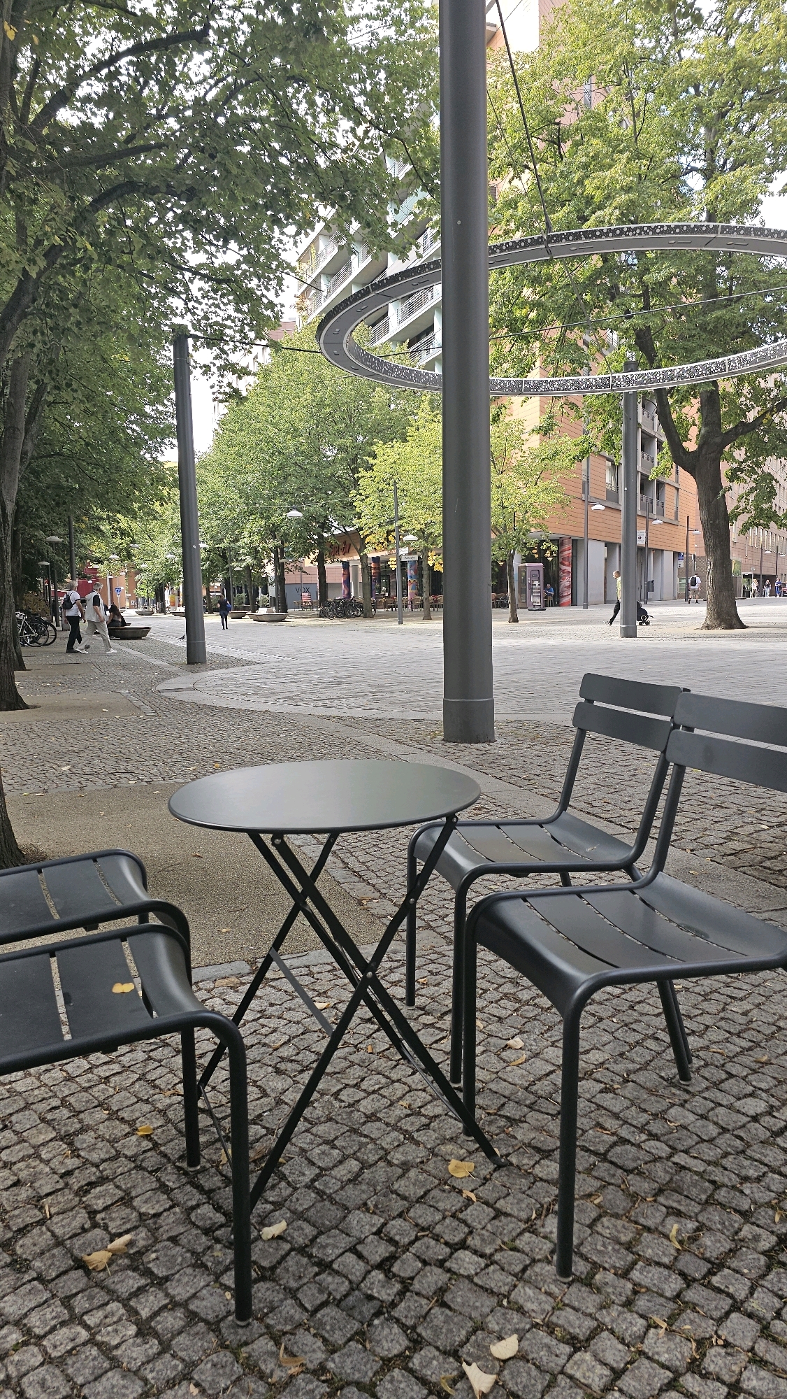 The image depicts an outdoor seating area in a European city, likely Berlin, Germany, characterized by cobblestone paving and mature trees. In the foreground, a dark grey, round, folding table is positioned with two matching chairs behind it. The chairs are made of a dark metal, with slatted seats and backs. The table has a thin, cross-shaped leg structure. Fallen leaves are scattered on the cobblestones around the furniture.
The background reveals a pedestrian plaza with more trees lining the sides. In the mid-ground, several people are visible. On the left, two individuals are walking away from the camera, one wearing a white shirt and the other in lighter clothing. Further back, a person in dark clothing walks towards the center of the plaza. Beyond the plaza, modern buildings with balconies and facades of brick and white elements are visible. A distinctive circular, illuminated art installation is suspended in the air, featuring a metallic ring with decorative lights. Street lamps and bicycles parked near the buildings are also present. The overall impression is a public space designed for relaxation and pedestrian circulation, possibly a park or a plaza within an urban environment.