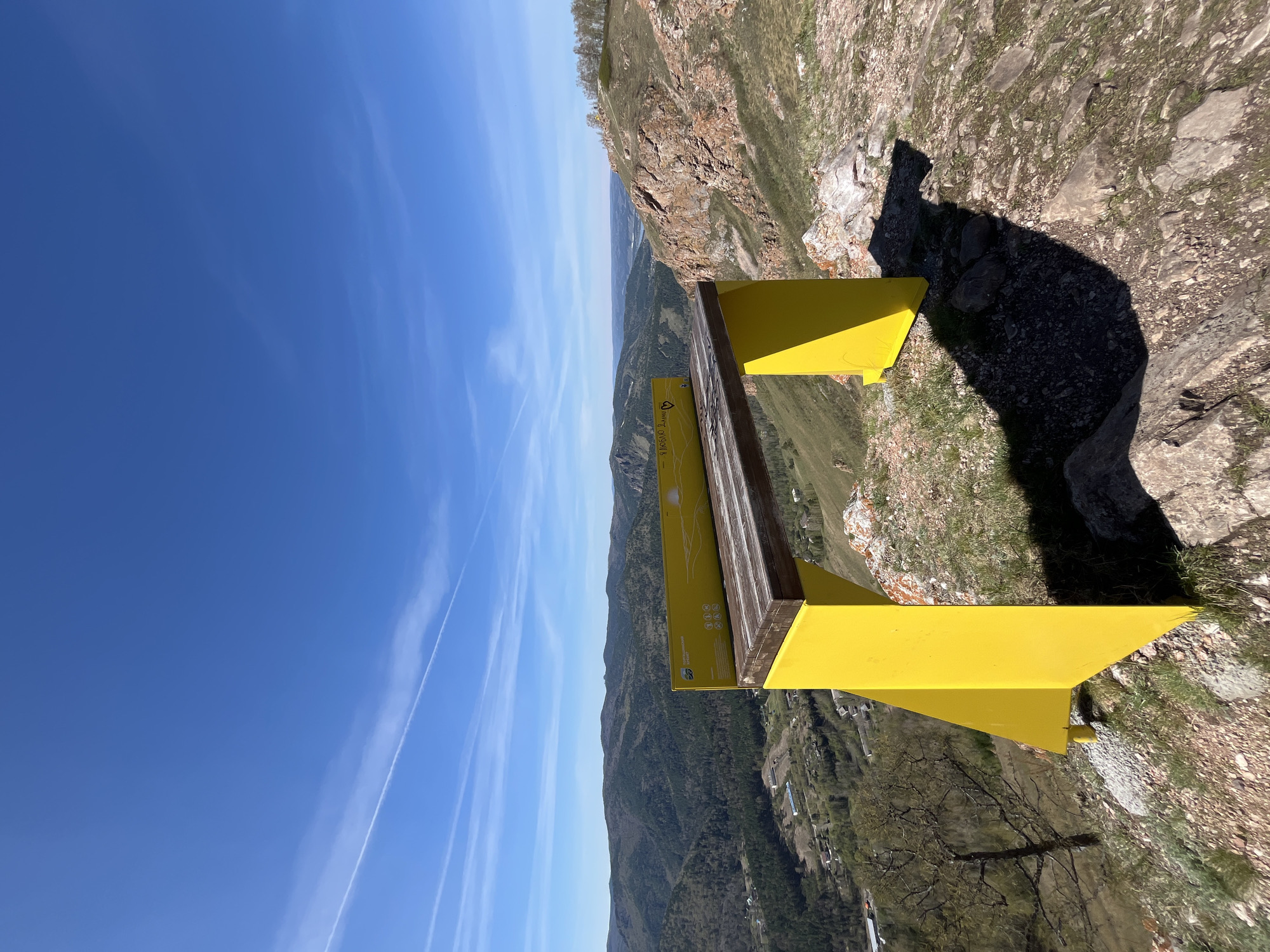 From a high vantage point overlooking a valley with a distant mountain range and a blue sky above, a bright yellow metal structure, likely an observation platform or viewpoint, stands on a rocky cliff. The structure has a wooden top and informational plaques, suggesting it is a tourist location. The landscape consists of verdant hills and valleys, punctuated by scattered trees and possibly small settlements. The sky is clear with faint contrails of high-flying aircraft. This location is situated in the city of Krasnoyarsk, Russia.
