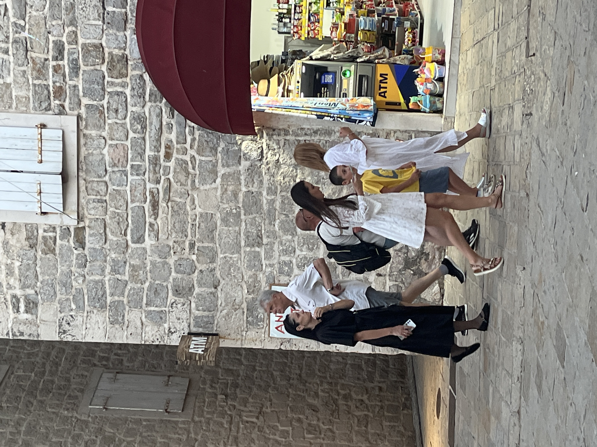 The image captures a street scene in Budva, Montenegro, focusing on a group of people walking past a store with an ATM. In the foreground, a woman in a black dress walks with a smartphone in her hand, followed by a man in a white shirt and gray shorts. To their right, a woman in a white lace dress walks alongside a young boy in a yellow t-shirt and blue shorts, followed by another woman in a white dress. Behind them, an ATM is visible outside a store filled with merchandise. The buildings are constructed of stone, and the street is paved with cobblestones. The scene is set outdoors, with natural lighting.