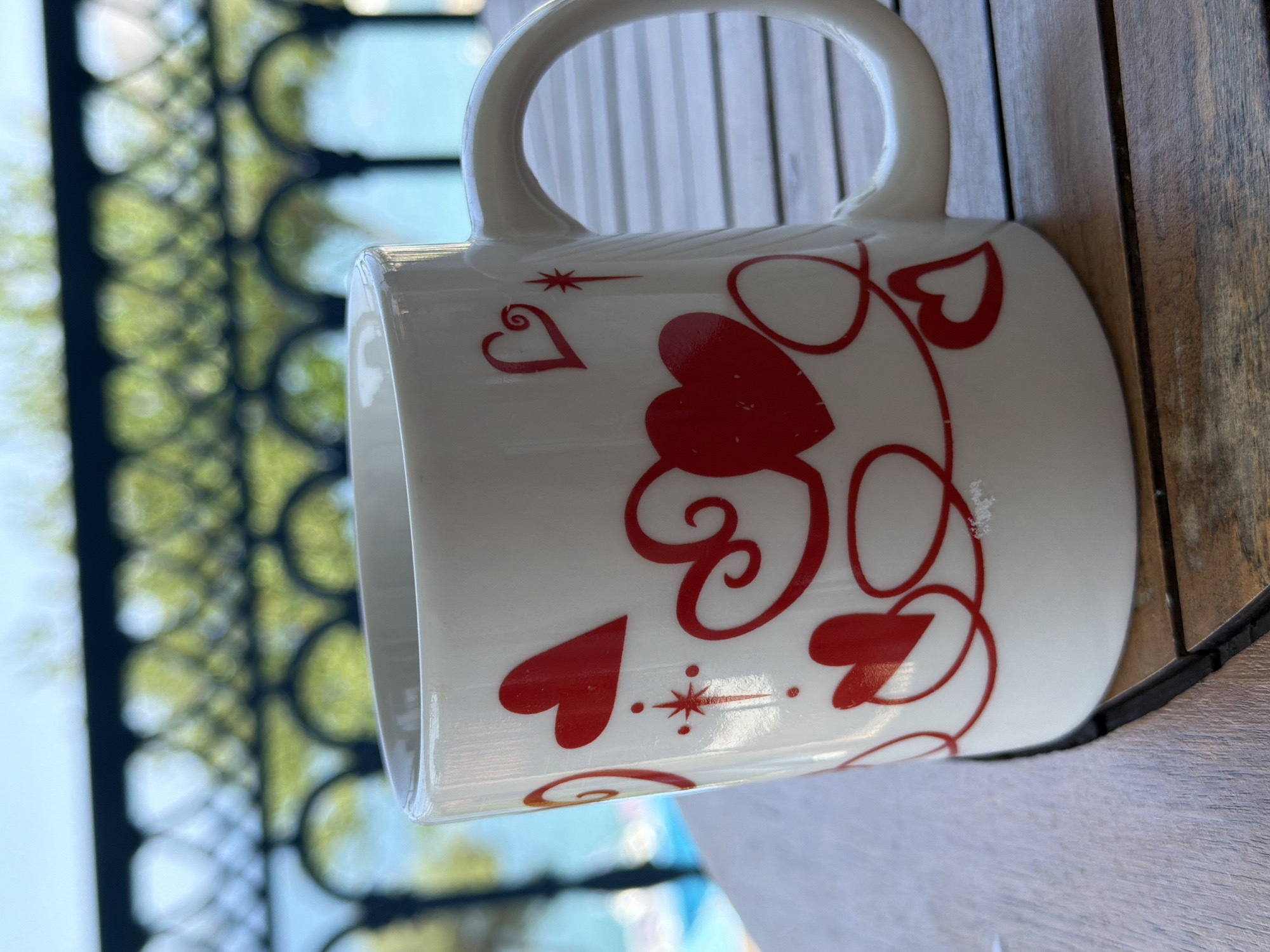 A close-up shot shows a white ceramic mug with a red heart design, placed on a wooden table outdoors. The background is blurred, revealing a dark railing and lush green foliage, suggesting an elevated outdoor setting such as a balcony or terrace, possibly in Municipal Unit of Alyzia, Greece. Beyond the greenery, a hint of blue water and sky is visible, possibly indicating a seaside location. The mug is the primary subject, with its decorative elements and placement on the table being the focus.
