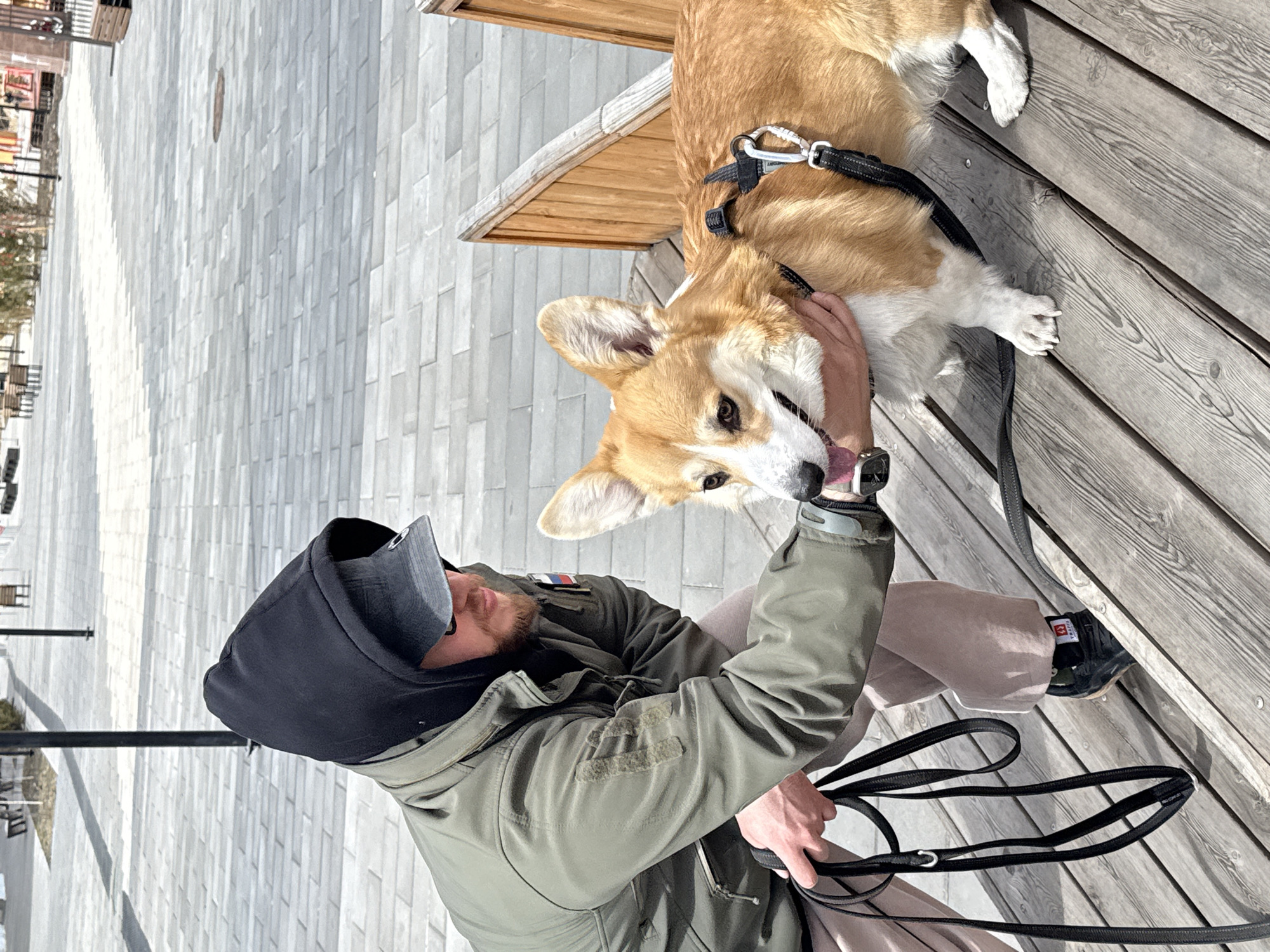 A medium, vertically oriented shot depicts a man interacting with a corgi dog. The man is on the left, wearing a dark hoodie under a green jacket and light tan pants. A baseball cap shades his face. He is kneeling, holding the corgi’s face with his left hand, and holding two black dog leashes with his right. The corgi is on the right, standing on a wooden boardwalk. The dog is light brown with white patches. In the background, a paved area is visible with street lights and building structures. The location is Borzya, Russia.