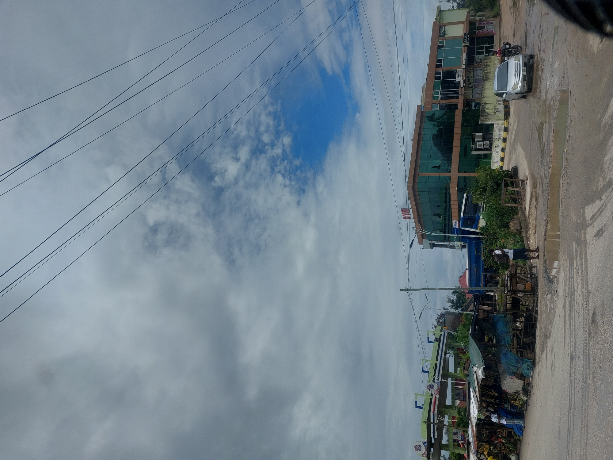 The scene depicts a street in Dar es-Salaam, Tanzania. A multi-story building with green glass windows stands alongside a street where a silver car and a motorcycle are parked. Adjacent to this, a roadside market is present, with individuals visible near stalls and a truck parked nearby. Above, a cloudy sky is intersected by power lines. The road surface is wet, suggesting recent rain.