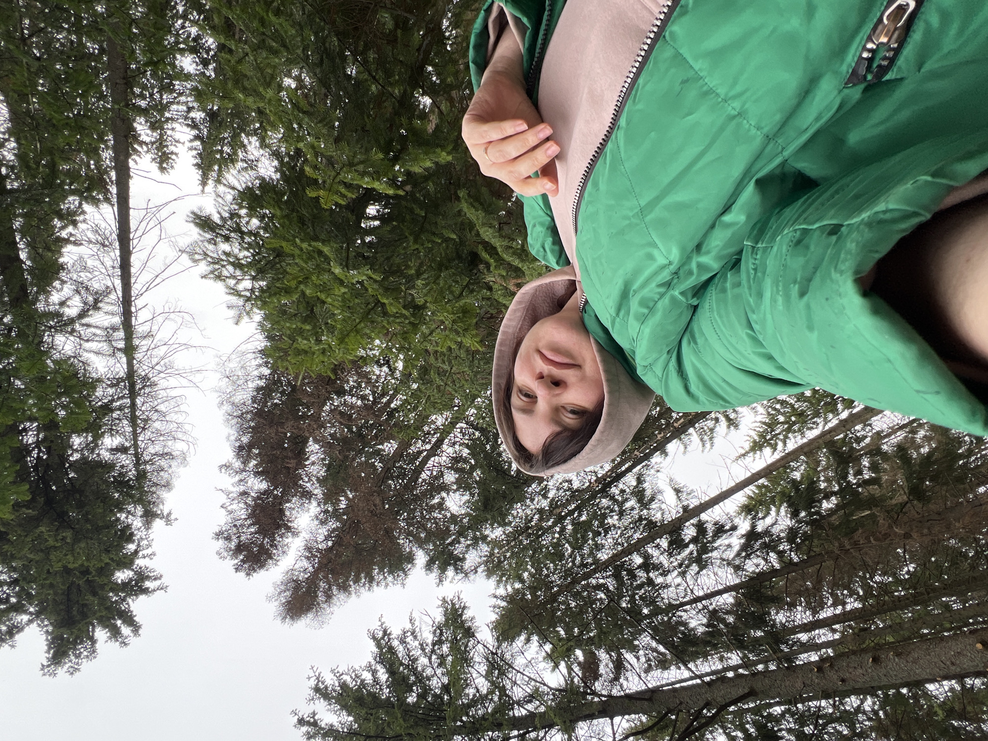 A young woman stands in a wooded area with evergreen trees. She wears a green jacket over a light pink hooded sweatshirt. The trees form a canopy overhead against a gray sky. The location is Bol'shakino, Russia.