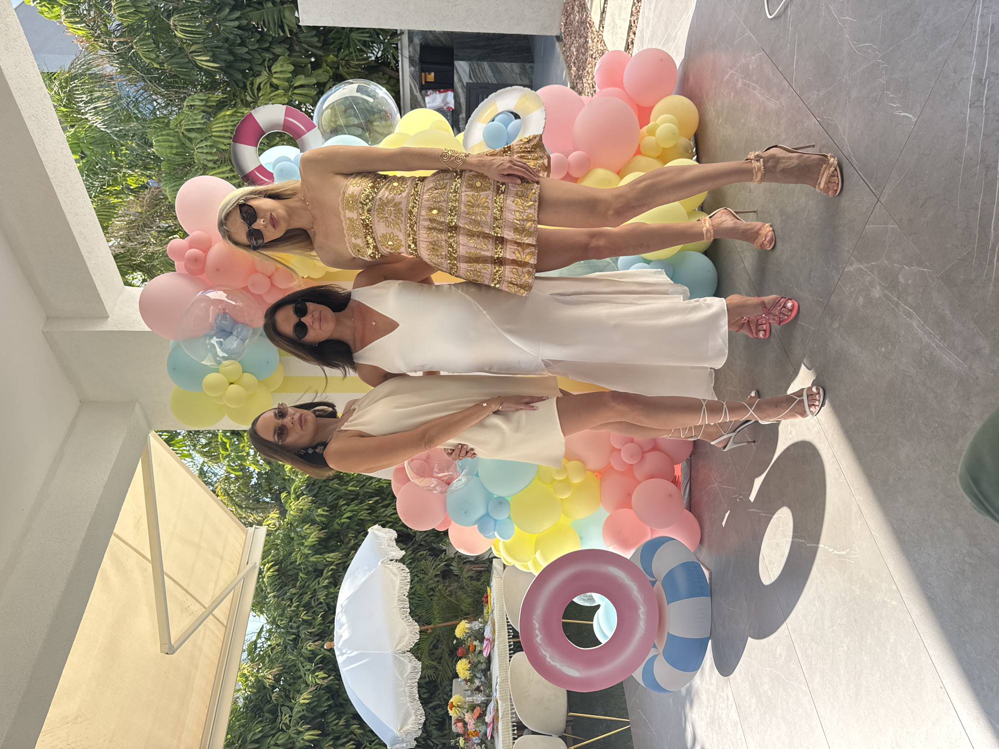 Three women pose for a photograph in an outdoor setting. The woman on the left wears a cream-colored slip dress and white strappy heels. The woman in the center wears a white slip dress and pink strappy sandals. The woman on the right wears a pink and gold sequined mini-dress and nude strappy heels. Behind them is a balloon arch in pastel colors, including pink, yellow, and blue, with some clear balloons containing smaller balloons. To the left of the women, a table is set with flowers, plates, and chairs, suggesting a party or event. In the foreground, two inflatable pool rings are stacked. The ground is paved with large gray tiles. The background shows lush green foliage and trees, with a glimpse of a building structure with a white awning. The lighting suggests a bright, sunny day. The location is identified as the city of Agios Tychona Community, Cyprus.