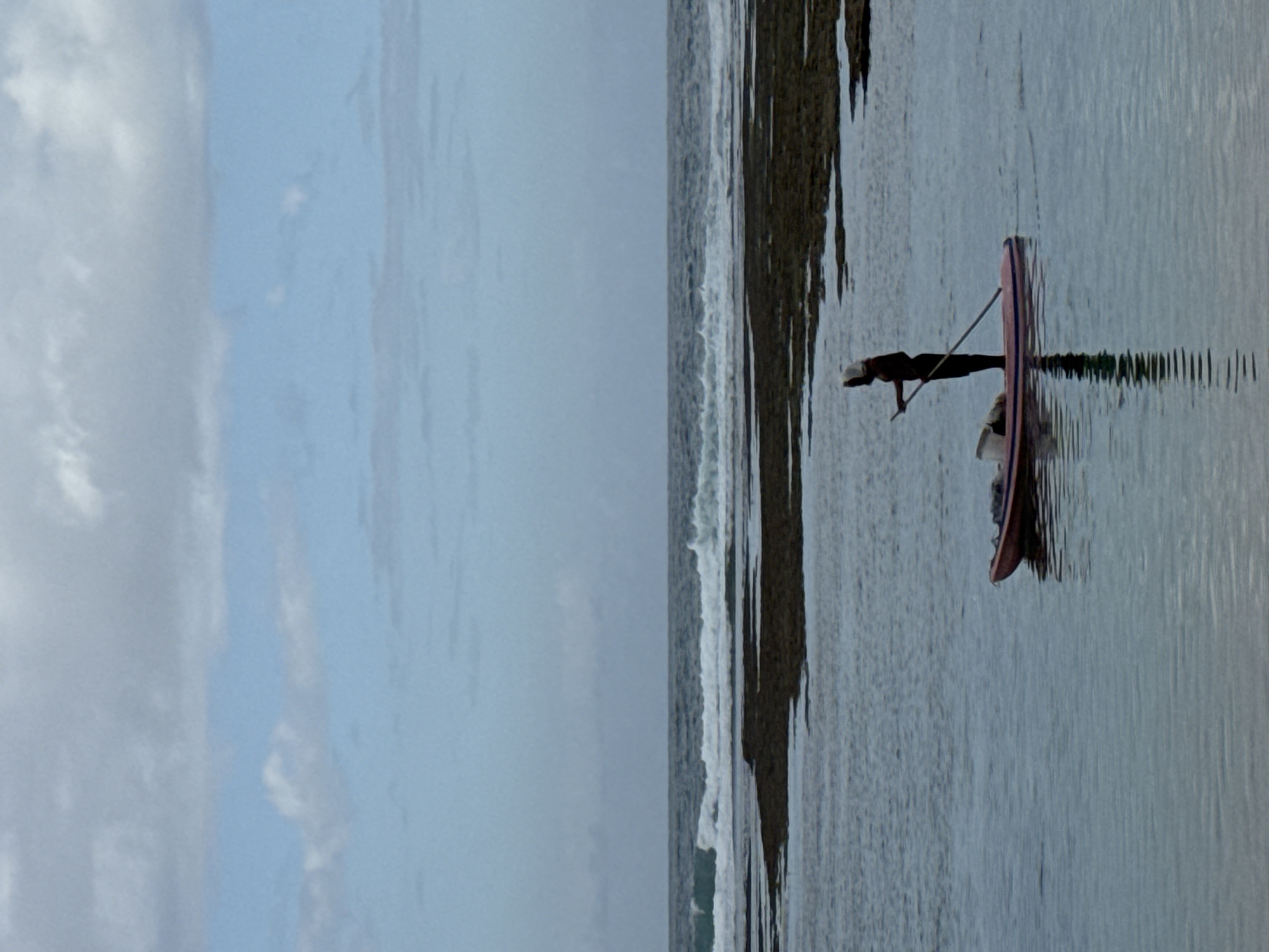 A lone figure stands on a paddleboard in shallow, reflective water. The figure, silhouetted against the muted sky, wears a light-colored hood. They are holding a paddle, its shaft extended across the board and into the water. The paddleboard is a pale pink with darker stripes along its sides, and appears to carry some items towards its front. Behind the paddleboarder, a dark, rocky outcrop breaks the surface of the water, and beyond that, gentle waves roll in from a calm, grey sea under a cloudy sky. The scene is calm and still, with the paddleboarder casting a sharp, dark reflection on the water's surface. The location is Kuta Selatan, Indonesia.