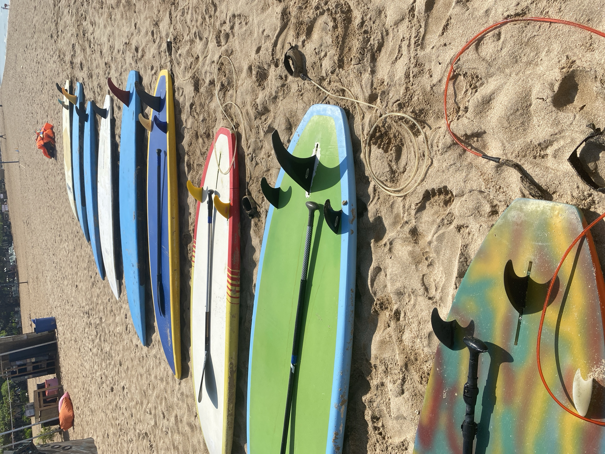 The image depicts a row of seven paddleboards lying on the sand, angled towards the upper right. From left to right, the boards are colored as follows: white, white, blue, blue, yellow, white with red stripes, green with blue stripes, and finally a multi-colored board. Each board is equipped with a paddle lying alongside it. Surrounding the boards is sand. In the background, a section of Nusa Dua, Indonesia, is visible. There is also an orange life jacket located in the background.