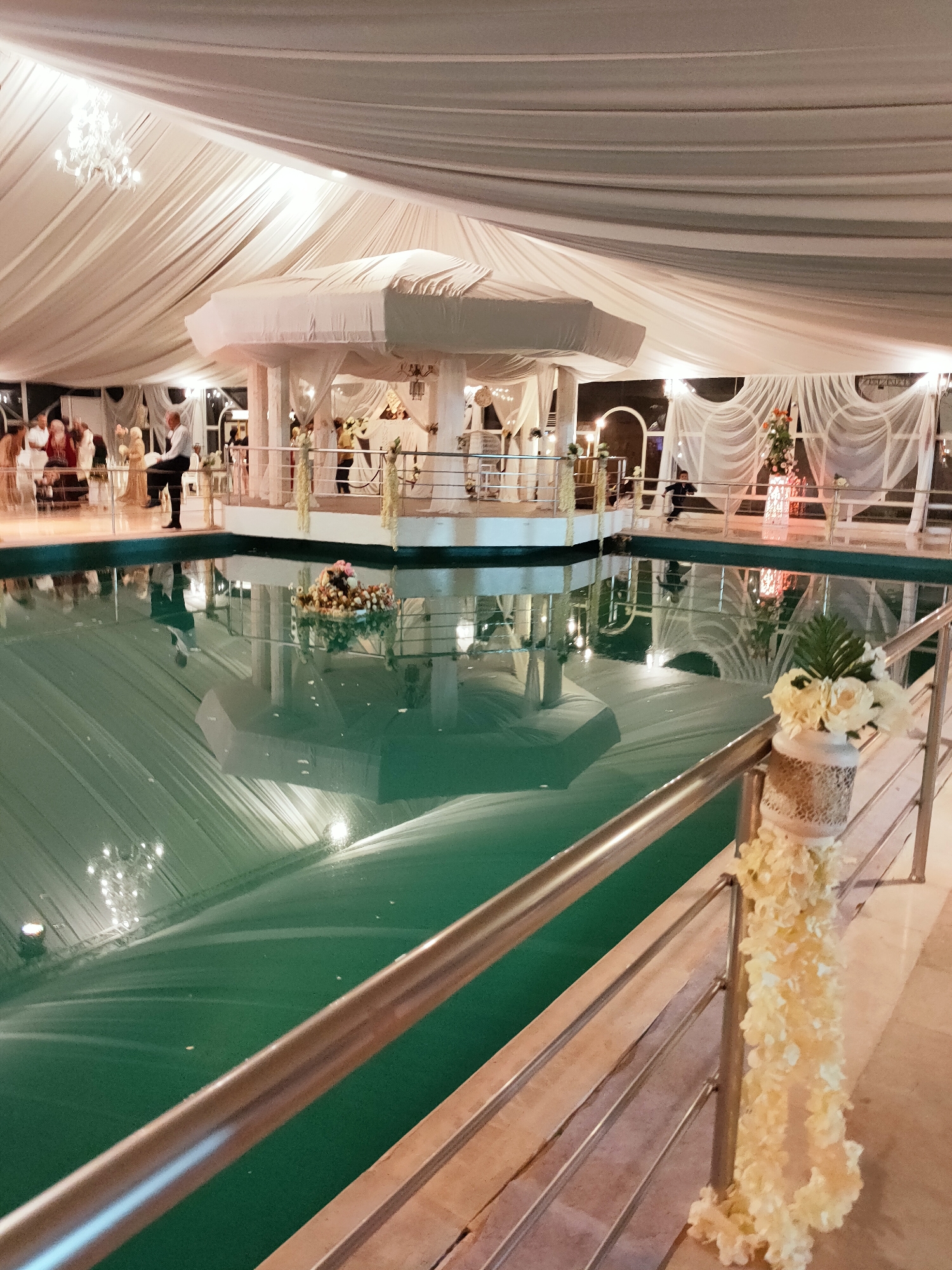 The image depicts an outdoor event venue, likely a wedding or party, in Mornag, Tunisia. The scene is dominated by a large, indoor swimming pool with green water, covered by a white draped tent. The tent's ceiling is adorned with chandeliers and flowing white fabric, creating a festive atmosphere.
In the background, a group of people are gathered near a white gazebo structure, which appears to be decorated with flowers and fabric. A man in a white shirt and black pants is standing by the pool's edge, facing the guests. There are also several floral arrangements placed throughout the venue, including a notable one on the railing in the foreground, consisting of white flowers in a decorative vase.
The lighting suggests it is evening, with artificial lights reflecting on the pool's surface and illuminating the tent and decorations. The overall impression is of a well-decorated, elegant outdoor event space.
