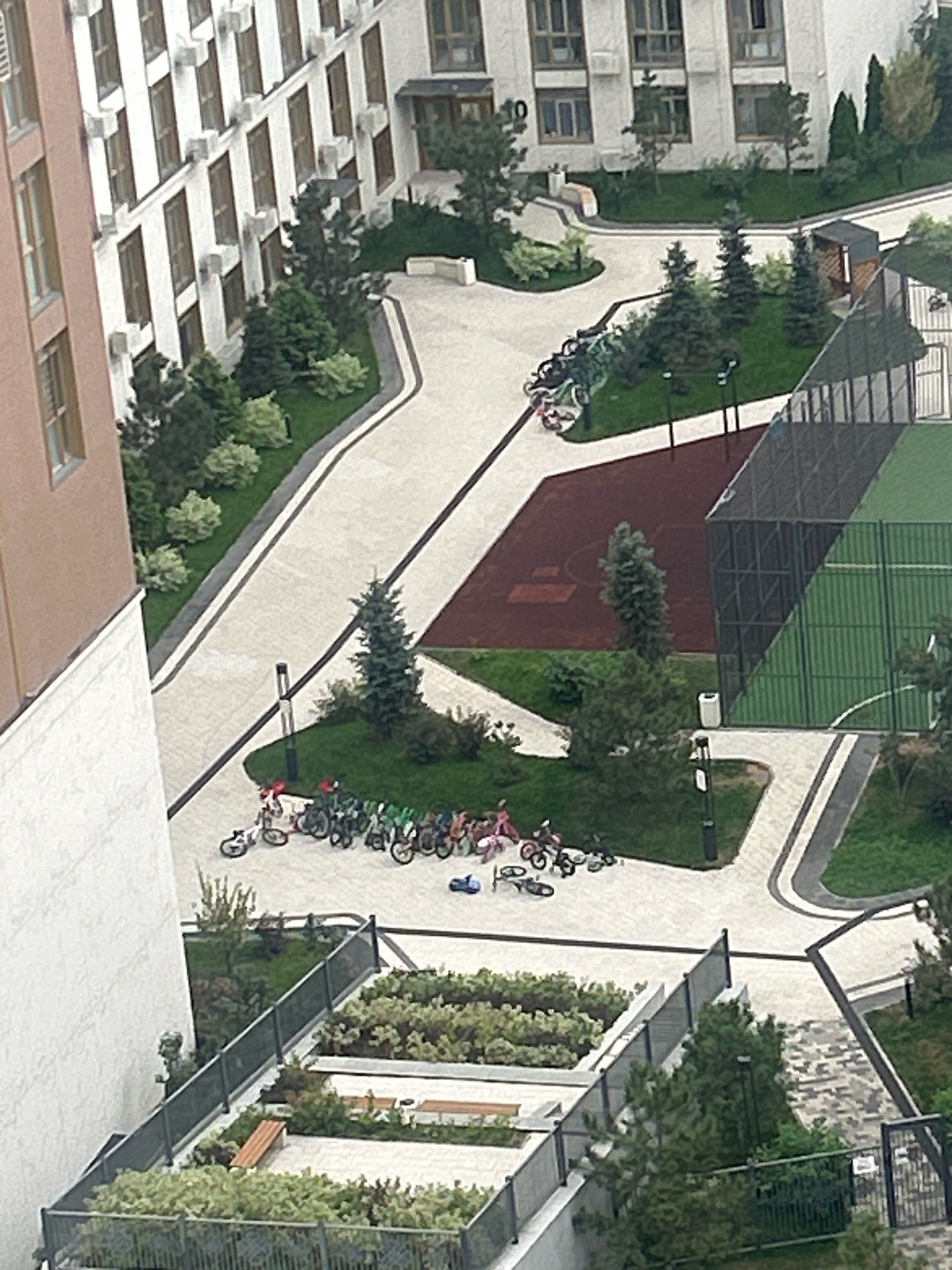 This is an aerial drone shot of a modern apartment complex in Almaty, Kazakhstan. The complex features a multi-story building with numerous windows and air conditioning units. In the foreground, there is a landscaped area with benches and greenery, enclosed by a fence. Adjacent to this, a children's playground area with several bicycles is visible. Further into the complex, a fenced sports court, possibly for basketball or tennis, is present, with a maroon-colored surface next to it. Trees and bushes are strategically planted throughout the complex, enhancing its aesthetic appeal. The sky is overcast, suggesting a cloudy day. There are no visible people in the immediate vicinity.