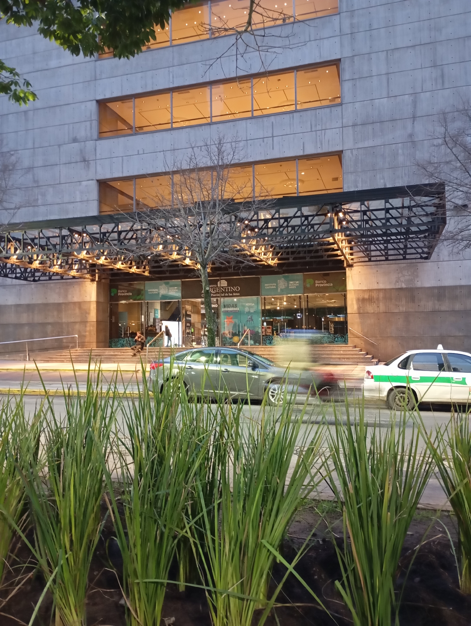 The image depicts the exterior of the "Centro Cultural de la Provincia de Buenos Aires - Teatro Argentino" located in La Plata, Argentina. The building features a modern facade with horizontal bands of windows illuminated from within. A dark metal structure with exposed lighting elements extends over the entrance area. In front of the building, a wide street is visible with two cars in motion, one appearing to be a taxi with green and white markings. Tall green grass lines the foreground, obscuring the lower portion of the street and the base of the building. Several individuals are present on the steps leading up to the entrance. The lighting suggests it is dusk or early evening.
