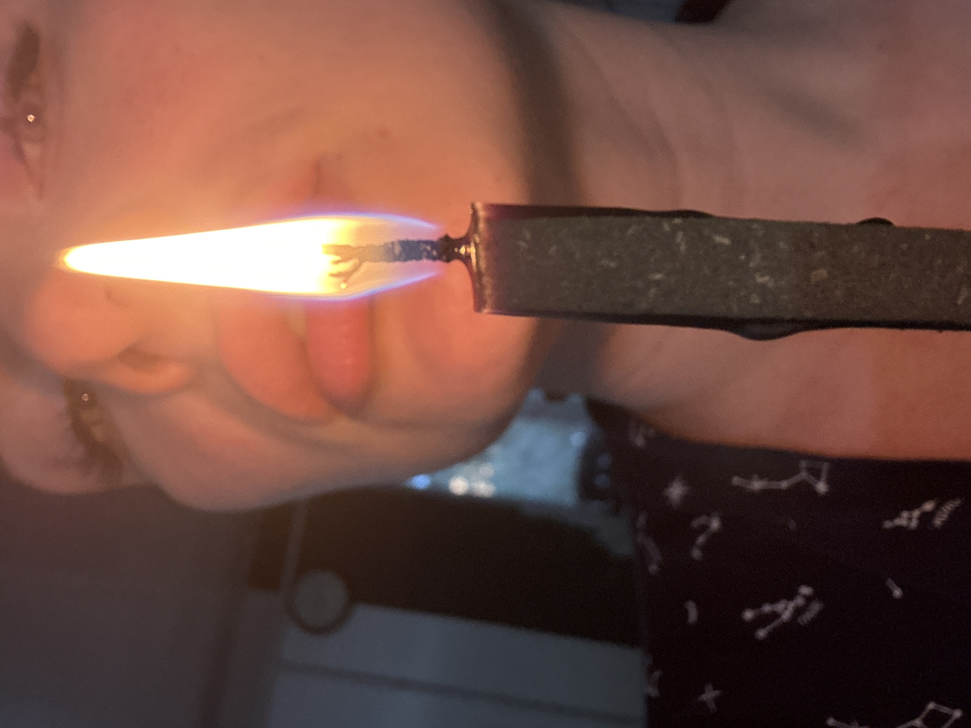 A person is holding a lit incense stick close to their face. The incense stick is dark gray and appears to be burning with a bright, elongated flame. The person is facing the camera, with a blurred background showing a dark surface with light reflections, and a garment patterned with constellation-like shapes. There is no indication of location.