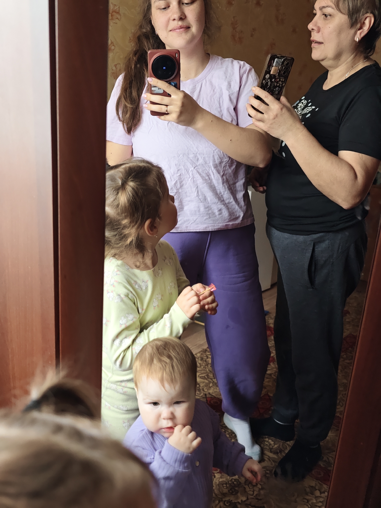 A mirror reflects two women holding phones, likely taking a picture or video of themselves and two young children. One woman wears a light purple t-shirt and dark purple sweatpants; the other wears a black t-shirt and black sweatpants. The older child wears light green pajamas with a bunny pattern, and the younger child wears a purple long-sleeve shirt. The scene appears to be indoors, with a rug on the floor. The location is Уральское, Russia.