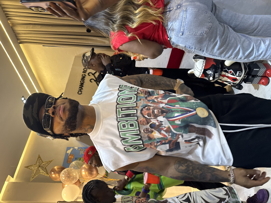 An indoor gathering features people, balloons, and decorative elements. A person in a white graphic t-shirt, camouflage hat, and sunglasses is prominently displayed. A woman in a red top and jeans is seen taking a photo with a phone. Other people are in the background, including one in a black jacket and camo hat. Decorations include a gold star and balloons. A black and white figure resembling a toy sits on a table. A sign reading "Changing Room" is visible.