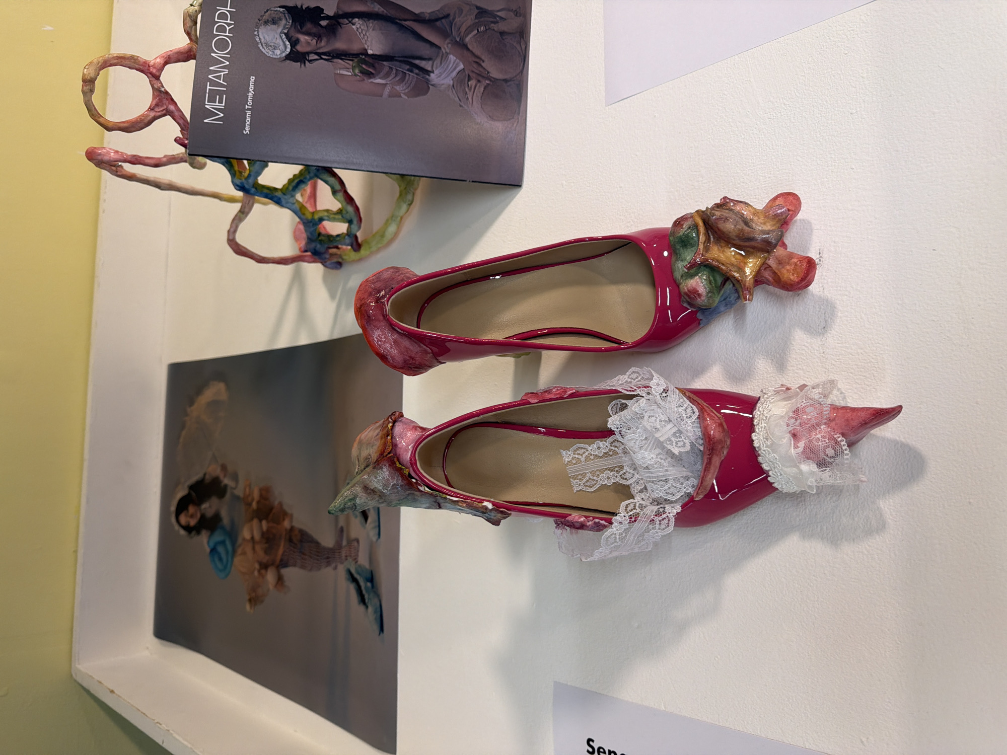 On a white wall, an art installation features two altered pink patent leather high-heeled shoes, embellished with ceramic or sculpted elements. The upper shoe has organic, anatomical-looking forms at the toe, while the lower shoe sports a lace trim and a pointed toe covered in a material resembling dried skin. A book titled "Metamorphosis" by Senami Tomiyama is displayed above the shoes, with a related sculpture of interwoven abstract shapes and a framed image depicting a person in elaborate, surreal garb to the left of the shoes. A partial label is visible beneath the lower shoe. The location of this art installation is Epsom and Ewell, United Kingdom.