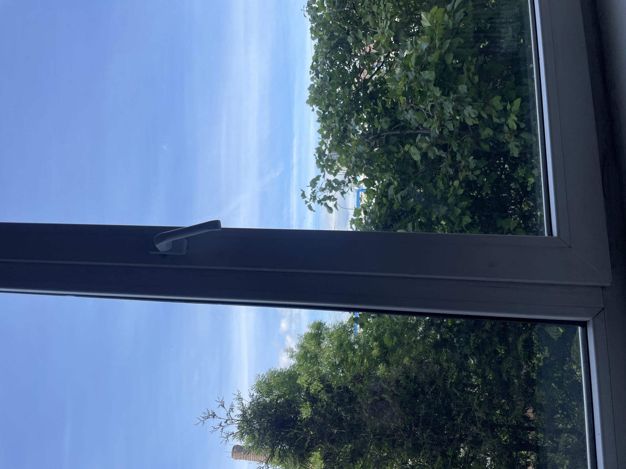 The image captures a clear, sunny daytime view looking out from a window in Saint Petersburg, Russia. A dark gray window frame, featuring a handle on its right side, partially encloses the scene.
The upper portion of the view is dominated by a vibrant, clear blue sky, showing only minimal, faint white atmospheric streaks. Below the sky, the mid-ground and foreground are filled with a dense, lush expanse of green tree foliage, indicating a healthy mix of deciduous trees. Through a break in the trees on the lower left, a reddish-brown brick chimney or industrial structure is partially visible.
In the distance, beyond the trees on the right side, a light blue, hazy horizon suggests the presence of a large body of water, likely the Gulf of Finland or a wide river. Along this distant horizon, faint outlines of buildings and structures are discernible, including a notable light blue object, possibly part of a building or infrastructure. The scene conveys a sense of serene natural beauty interspersed with distant urban elements under a bright sky. No visible text is present.