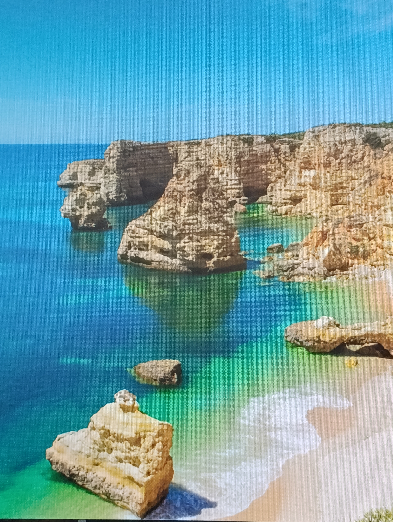 The image displays a coastal scene featuring a beach bordered by rugged cliffs and rock formations. The water transitions from a deep blue to a vibrant turquoise near the shore, suggesting varying depths. Waves gently lap against the sandy beach. Several large, yellowish-brown rock structures are scattered in the water near the beach and along the cliff line. The cliffs themselves appear to be composed of layered rock formations. The sky is a clear, light blue. The location is a beach in an unknown city.