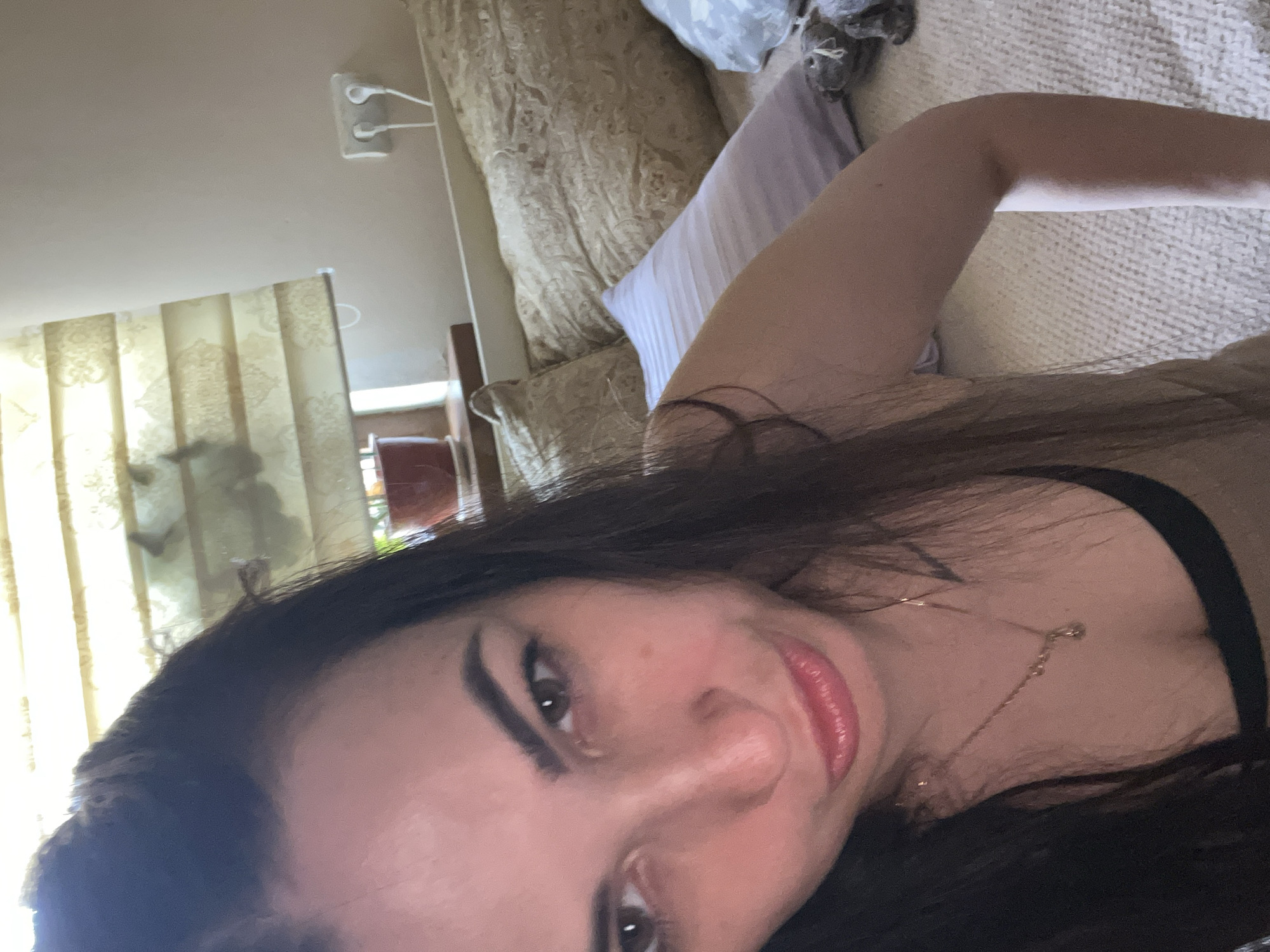 A woman is taking a selfie in a room. She has long dark hair and is wearing a tan top. She is sitting on a bed with pillows behind her. The room has blinds on the window and an electrical outlet on the wall.
