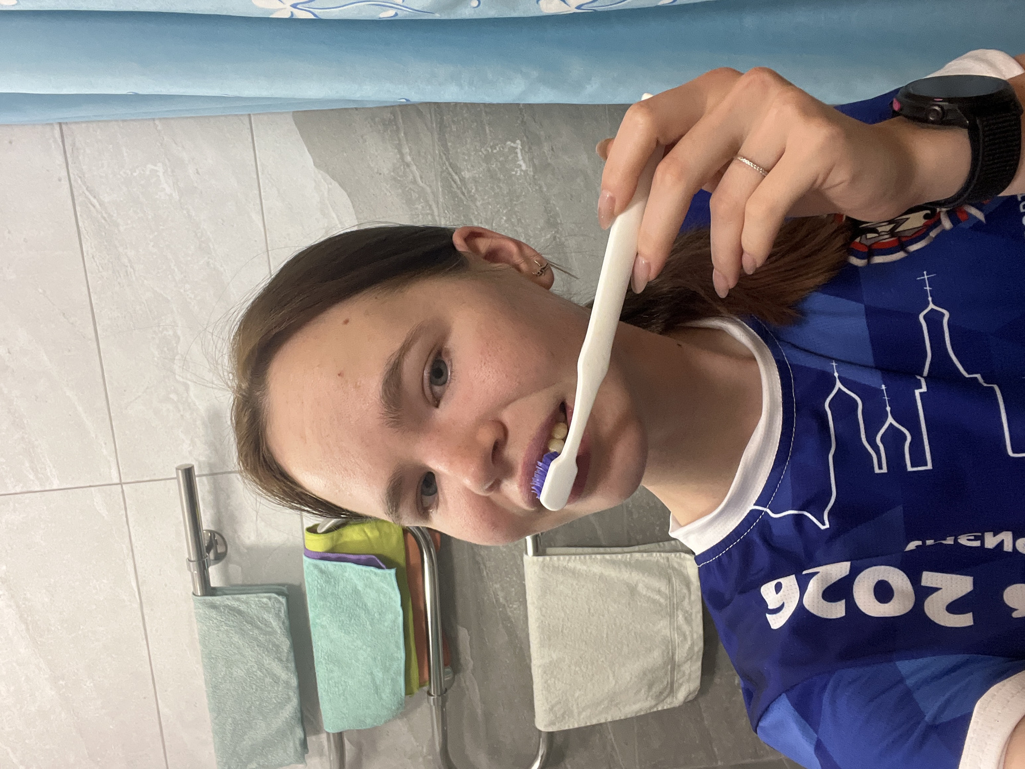 A young woman is in a bathroom, holding a toothbrush with blue bristles to her mouth. She is wearing a blue jersey with white trim and the year "2026" printed on it along with outlines of a building resembling the Cathedral of the Assumption in Perm, Russia. She also wears a black smartwatch on her left wrist. The bathroom has gray tile walls, a multi-towel rack with green, yellow, and purple towels, and a white towel hanging on a separate rack. A light blue covering is visible above the wall. The location is the city of Perm, Russia.