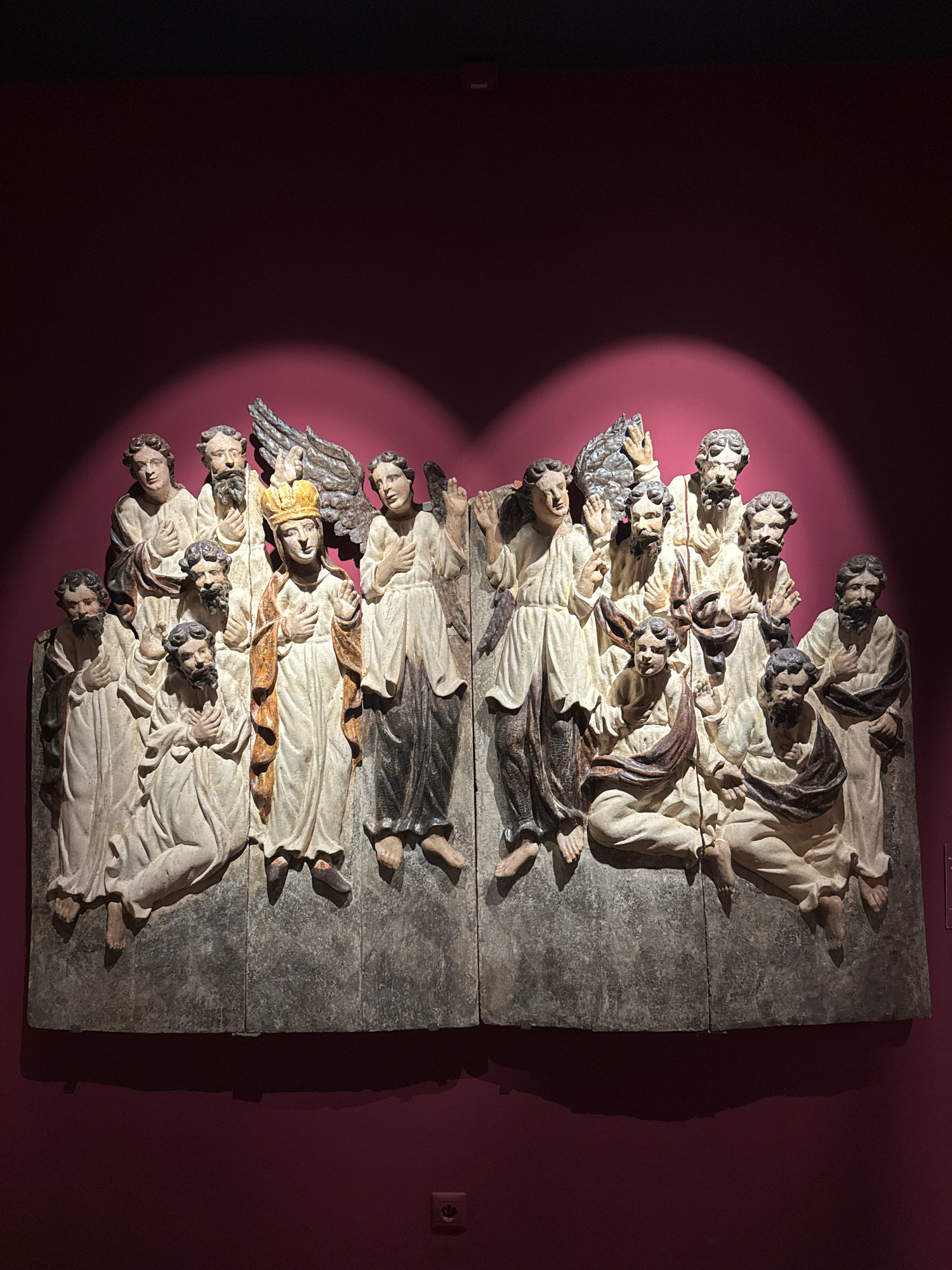 An overhead spotlight illuminates a large, weathered, carved relief depicting a group of figures, likely religious in nature, against a deep red wall. The relief is divided into two vertical sections. The left section features seven figures. In the center of this section stands a crowned figure wearing a golden and white robe, with one hand raised. To their left are two figures standing behind a kneeling figure, and to the right are three standing figures, one with a feathered wing. The right section displays six figures, with one standing figure on the far left, a group of three figures clustered in the center, and two more figures on the right. Some figures are seated and others stand, all adorned in draped robes. The figures appear to be made of wood and have a aged, somewhat worn appearance. The base of the relief is a dark, textured stone-like material. Below the relief, a prominent shadow is cast on the wall, and a small, light-colored electrical outlet is visible on the lower right side of the red wall.