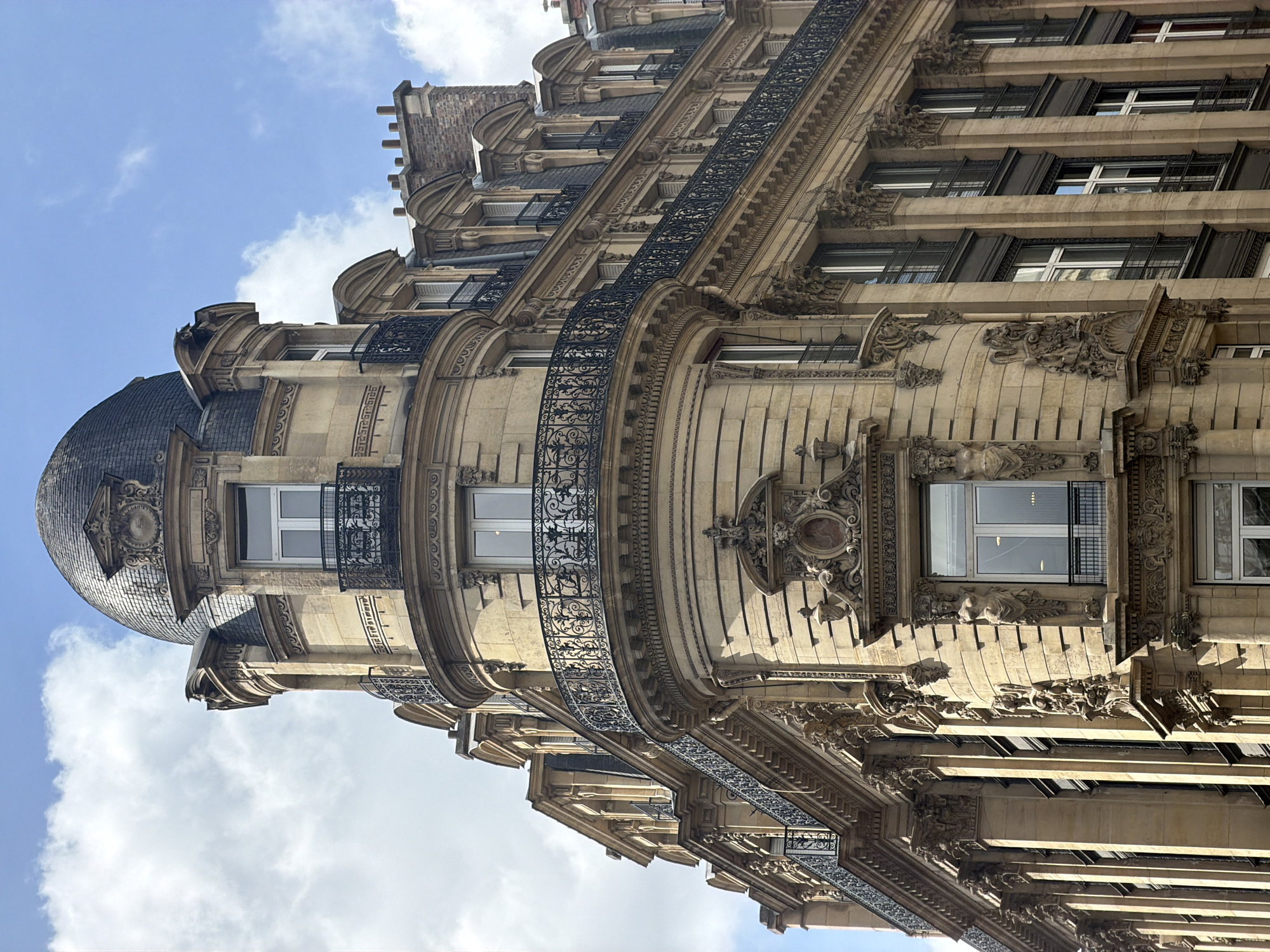 A grand, ornate building in Paris, France, is captured from a low-angle perspective, looking upwards. The impressive structure showcases classic Haussmannian or Beaux-Arts architectural styles, characterized by a prominent curved facade that forms a corner. A dark, slate-tiled dome or cupola crowns the upper corner of the building, adding a distinctive feature.
The facade is constructed from light-colored stone, adorned with multiple stories featuring continuous, elaborate wrought-iron balconies that trace the building's curves. Each floor displays rectangular windows, some with dark frames or exterior elements, and extensive sculptural details. These include intricately carved reliefs, decorative brackets, garlands, and ornate mouldings, which highlight the craftsmanship. Towards the lower right of the image, the facade reveals more intricate stone carvings, possibly including figural elements or caryatids. A section of red brickwork is visible at the top, likely part of an attic level or chimney structure.
The scene is brightly lit by natural daylight, casting shadows that accentuate the three-dimensional depth and rich textures of the stone. Above the building, a clear blue sky is visible, dotted with white, fluffy clouds. No people or text are present in the image.