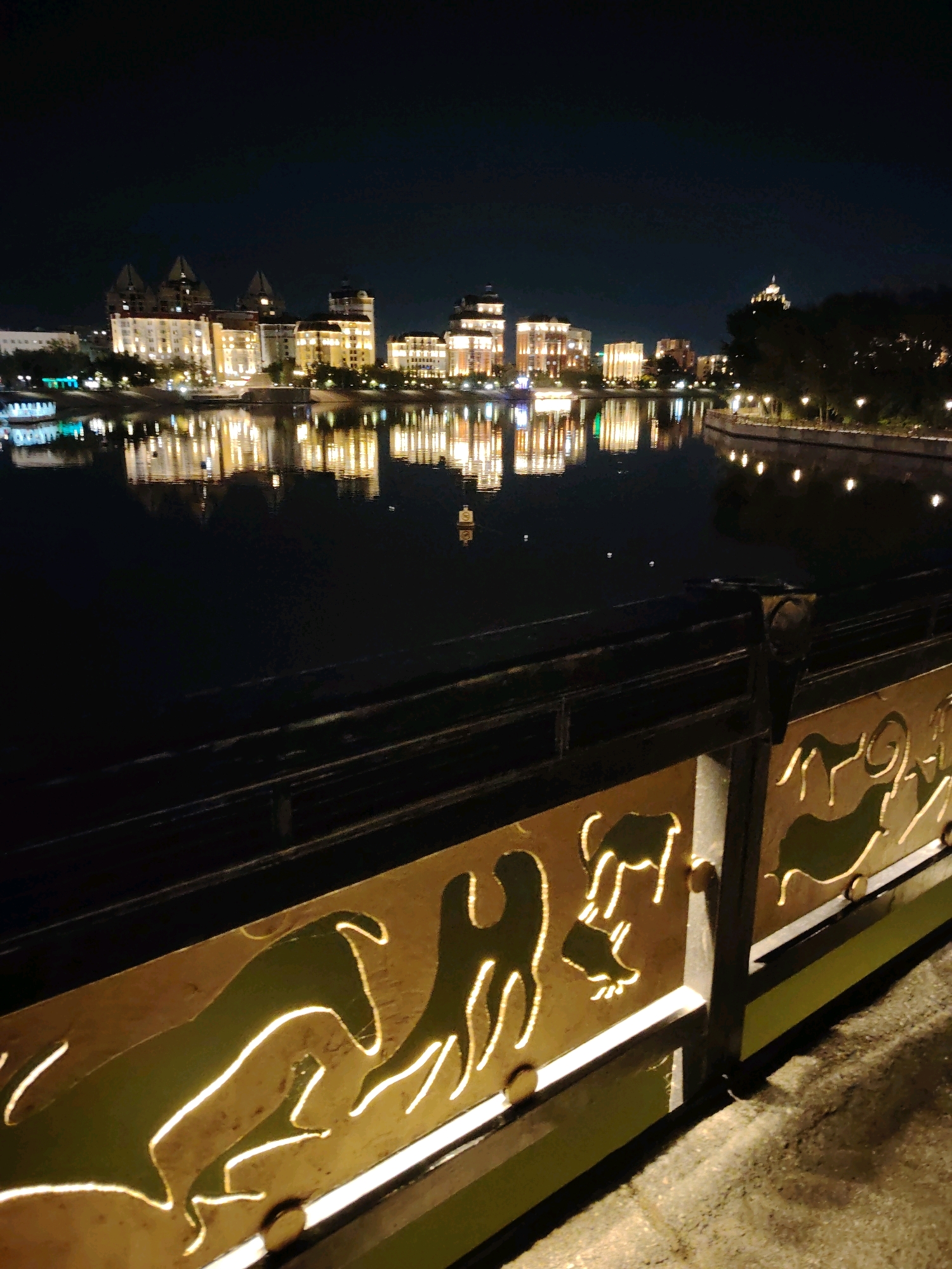 The image depicts a night scene along the Ishim River in Astana, Kazakhstan. In the background, illuminated buildings with distinctive architectural features line the riverbank, their lights reflecting on the dark water. The foreground features a decorative railing with an intricate design of animals, illuminated from behind, creating a striking contrast against the night. The railing appears to be made of metal with a golden finish, and the cut-out shapes allow light to shine through, casting shadows on the ground. The river itself is calm, with gentle ripples disturbing the reflections of the cityscape. The overall atmosphere is one of urban tranquility and illuminated beauty at night.