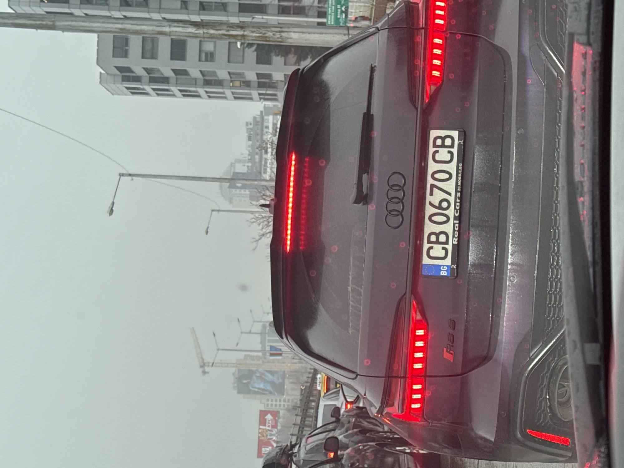 The image captures the rear of a dark-colored Audi RS6 sedan with a Bulgarian license plate "CB 0670 CB". The vehicle is positioned in heavy traffic in Sofia, Bulgaria. The backdrop shows a rainy cityscape with buildings, streetlights, a construction crane, and a billboard featuring a KFC advertisement. The photograph was likely taken from inside another vehicle, looking out through a slightly wet or smeared windshield. The general atmosphere is overcast and gray due to the weather.
