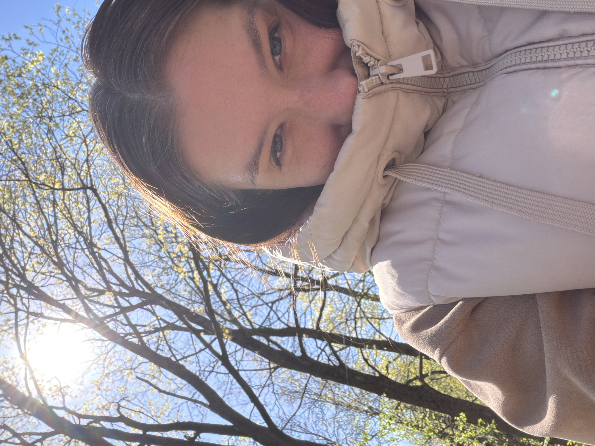 A close-up, slightly tilted, eye-level shot depicts a person partially obscured by a light beige, possibly insulated jacket, against a backdrop of bare tree branches and a bright blue sky. The sun is visible in the lower left, creating lens flare. The person's eyes are visible above the jacket's collar. The location, Tokkari, Russia, is not deducible from this photograph.