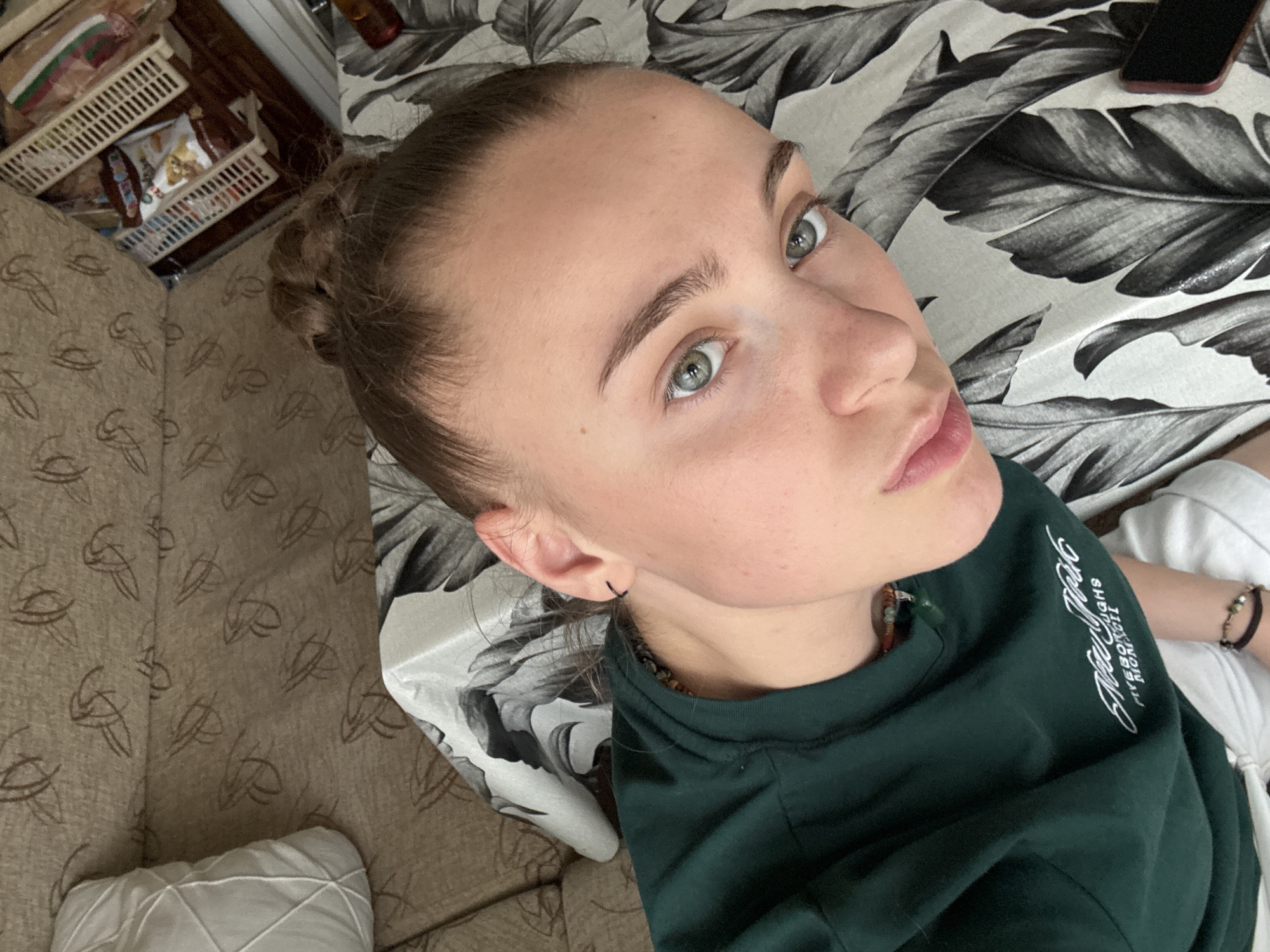The image is a close-up, overhead shot of a young woman lying down. She has her hair in a bun and is wearing a dark green long-sleeved shirt with white text on the front, which appears to say "New York Five Boroughs". She is wearing a small hoop earring in her left ear and a beaded necklace. Her eyes are green and her skin is fair. She is lying on a patterned fabric, possibly a sofa or bedspread, with large black and white leaf designs. To the left of the woman, a light-colored pillow with a quilted pattern is visible. In the upper right background, a shelf unit with various items, including baskets of snacks, can be seen. The lighting suggests an indoor setting. There are no visible landmarks or elements that would help pinpoint the location to Gabrovo, Bulgaria.