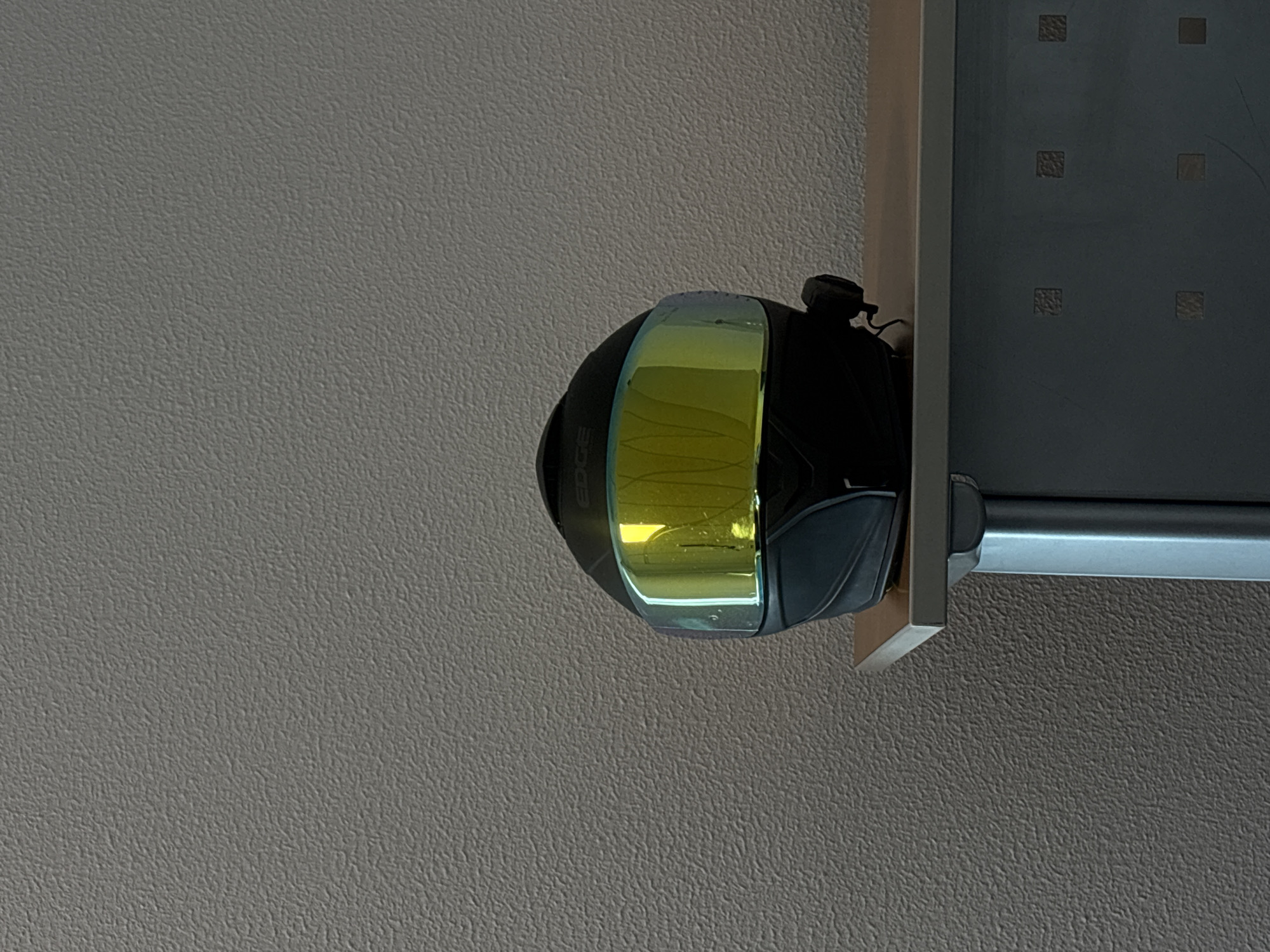 A motorcycle helmet with a yellow visor is hanging on a silver-colored towel rack mounted to a wall. The wall appears to be painted in a neutral color with a textured surface. The background includes a patterned, dark-colored surface possibly a door or a cabinet, adjacent to the helmet. The environment suggests an indoor setting, such as a room or a closet. There are no people visible in the image. The location, Puebla, Mexico, is not discernible from the visual information provided.